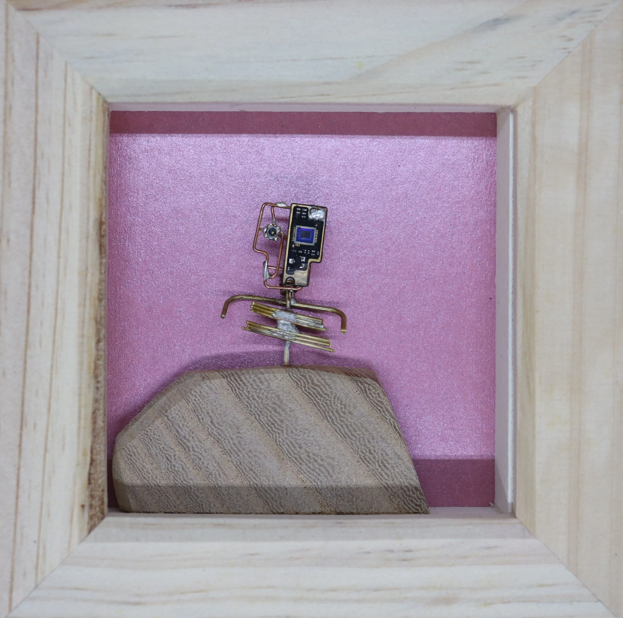 A miniature wooden frame with pink background, featuring a small rock, a tiny electronic circuit, and several miniature wire hangers arranged to resemble a person inside the box.