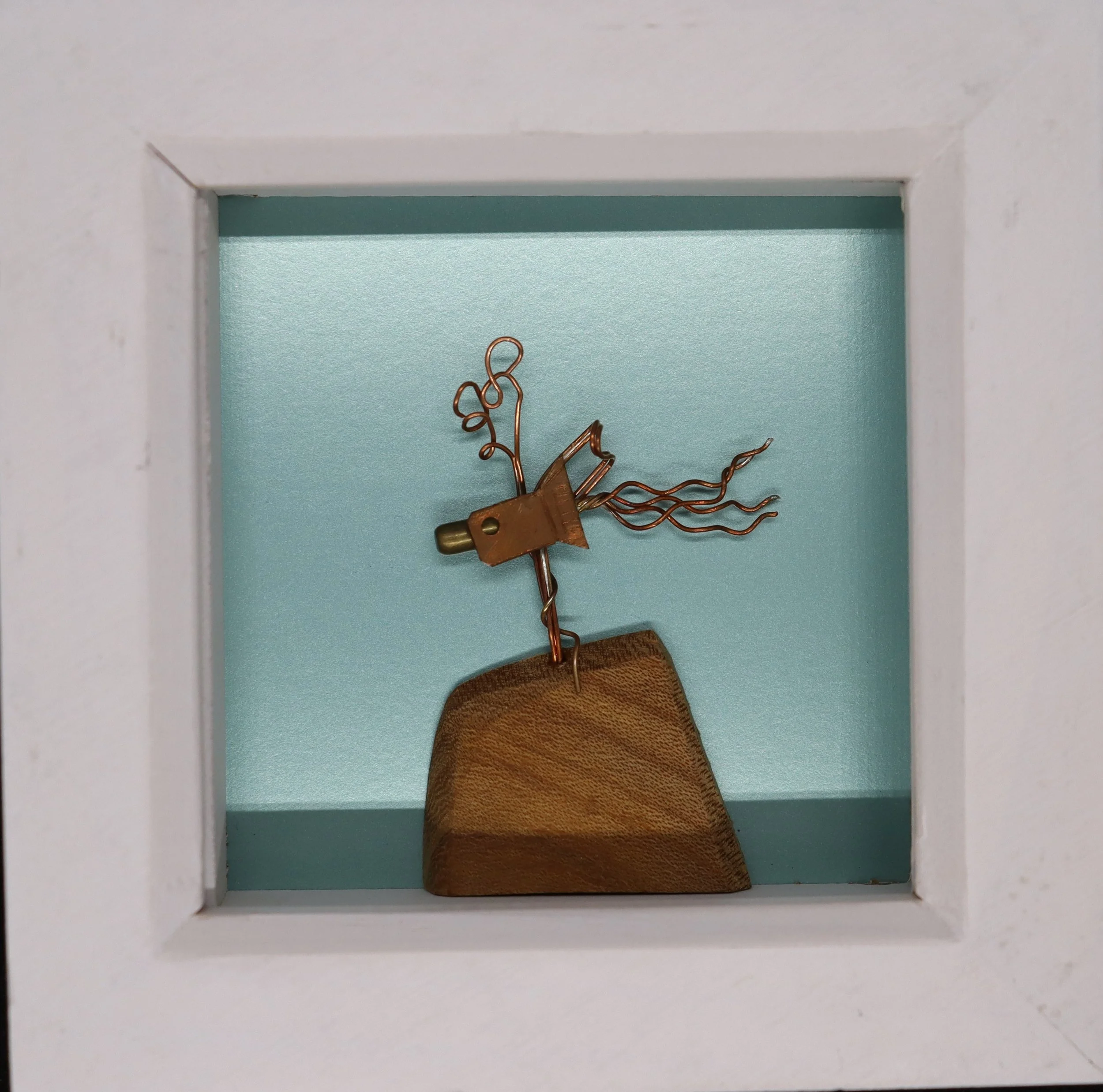 Small sculpture of a fairy with wire wings and hair, perched on a wooden block, enclosed in a white shadow box.