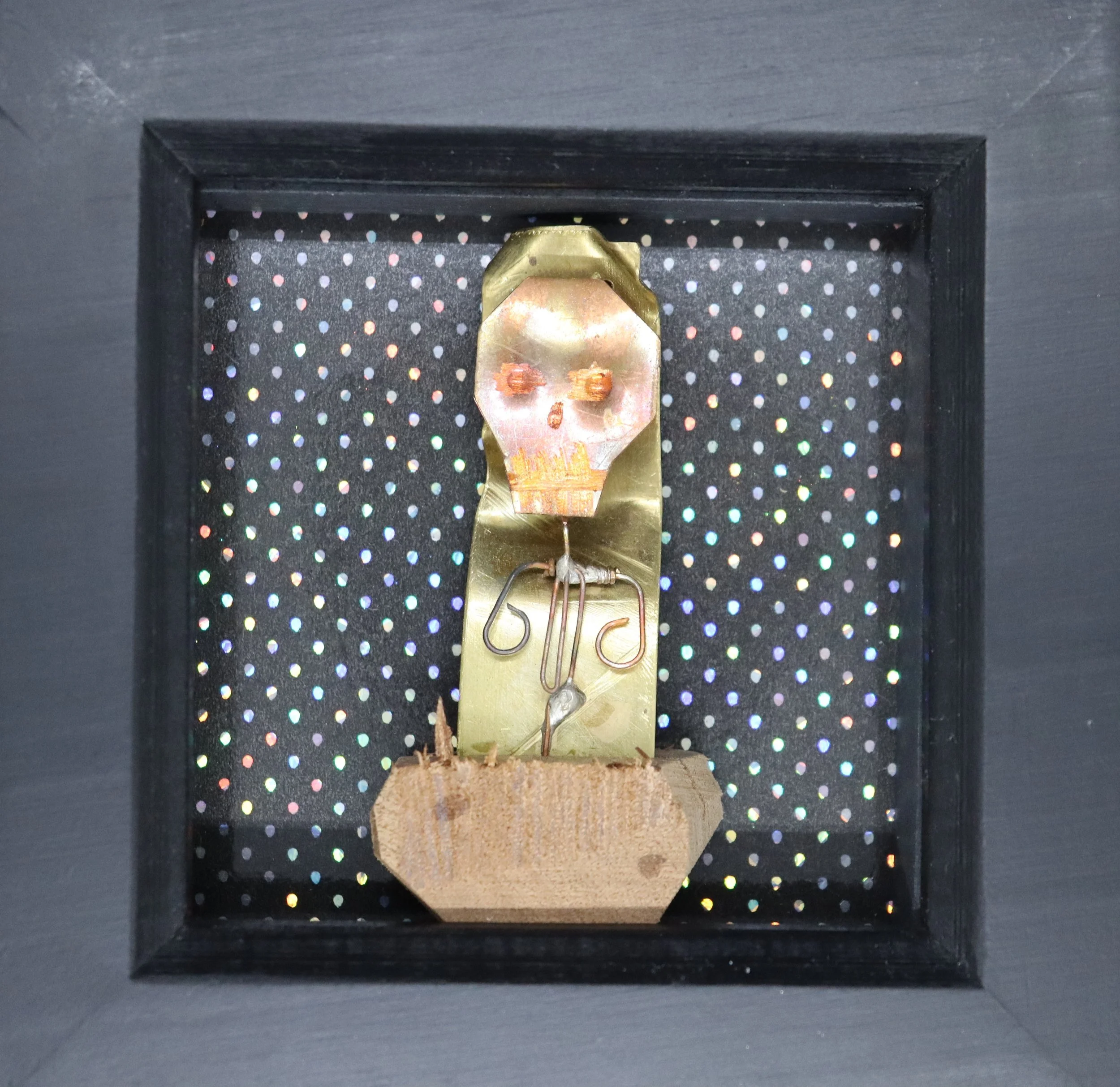 Close-up of an antique clock movement with a skull face drawing, mounted inside a black display case with a dotted iridescent background.