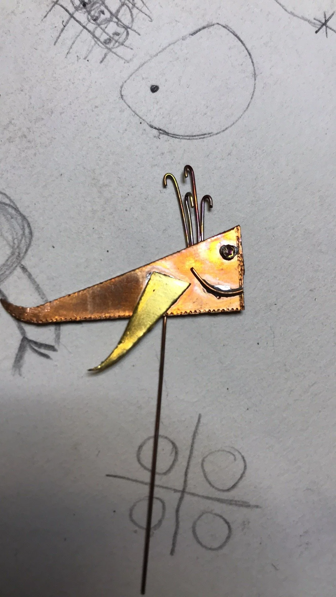 A craft project of a smiling animal, possibly a dog, made from metal and paper, on a background with sketches and tic-tac-toe drawing.