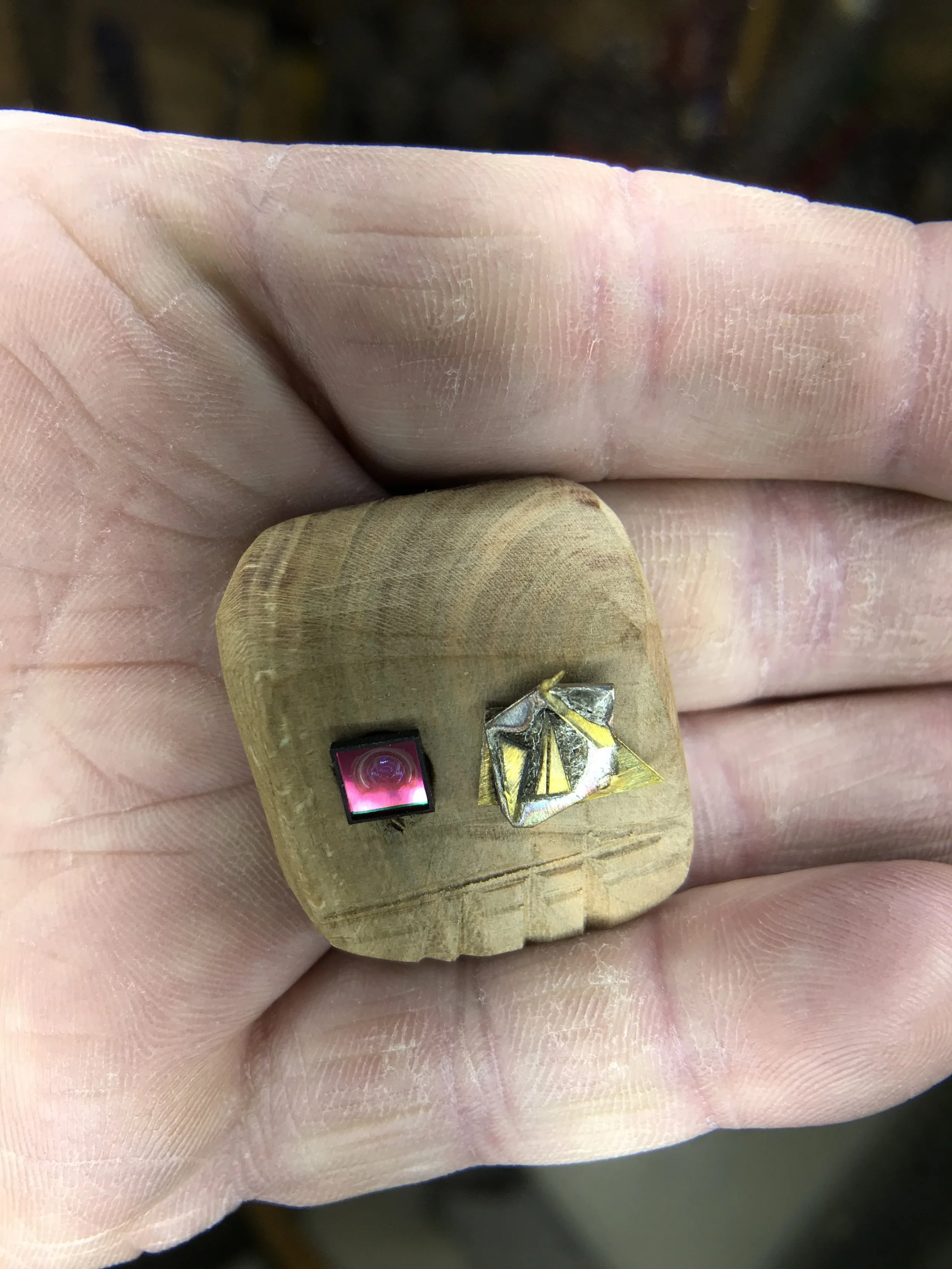 A hand holding a small wooden block with two tiny objects on it: a black square with a pink spiral design, and a metallic object with gold and silver parts.