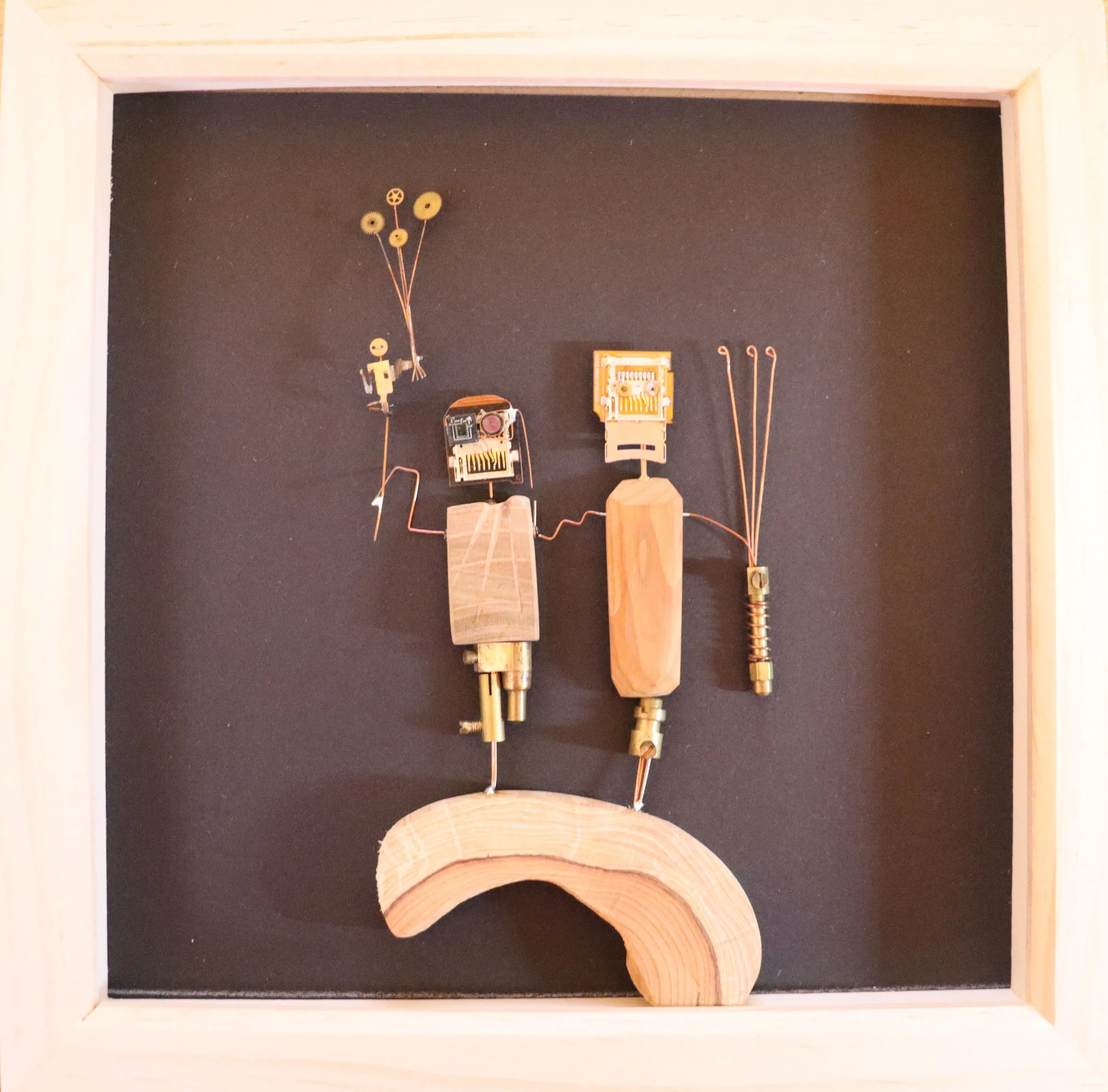 Mixed media artwork of a humanoid figure made from wood, metal, and electronic parts, standing on a curved wooden base, with wire and small objects forming hair, arms, and accessories, set in a wooden frame.