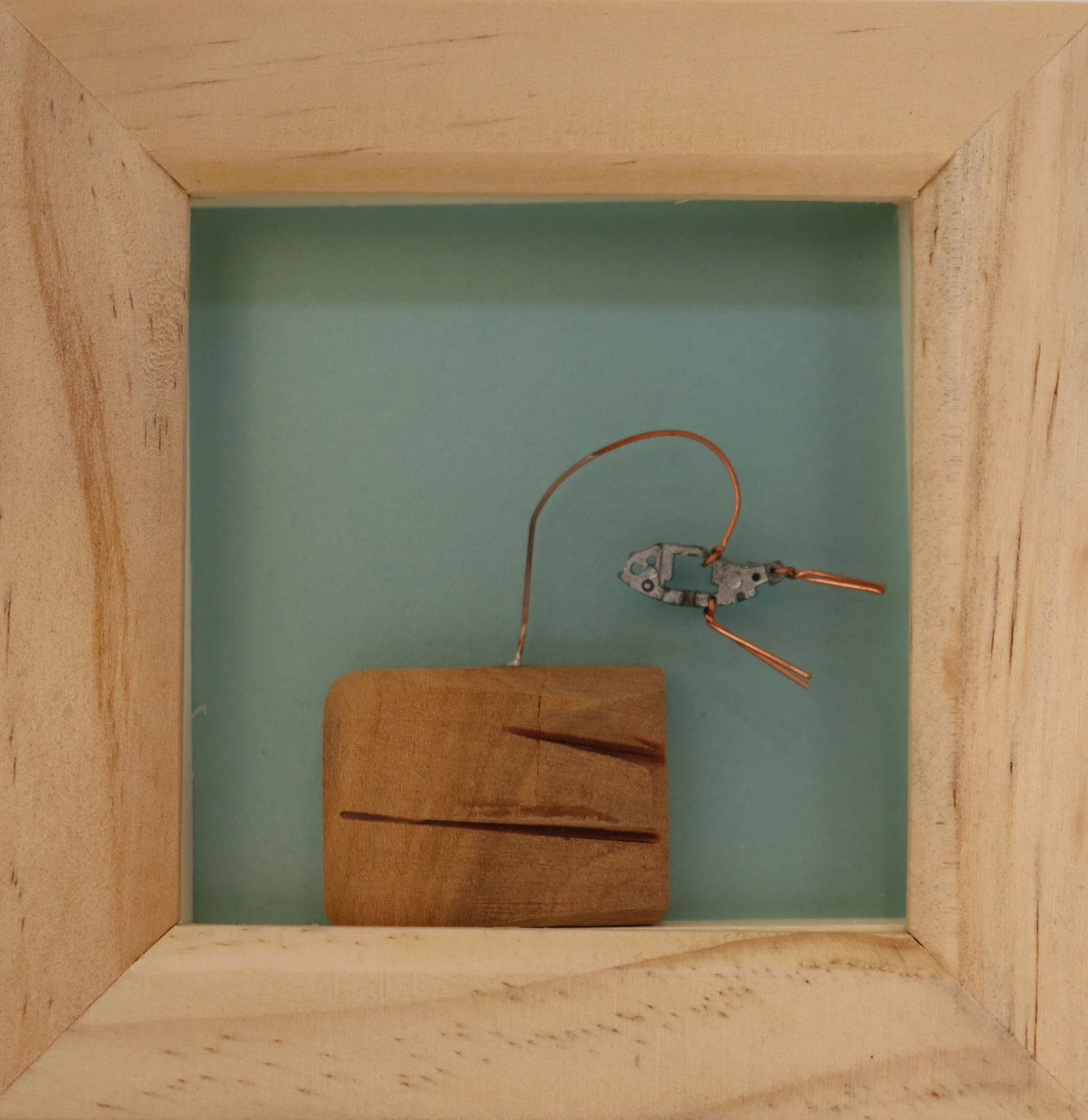 Inside a wooden box, a small mechanical part with a metal wire and copper wires attached, resting on a wooden block with visible cracks.