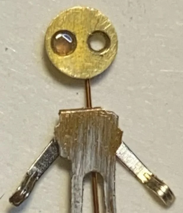 A small humanoid figure made from various hardware parts, with a round brass washer as the head with two holes resembling eyes, a metal clip for the body, and bent metal pieces as arms and legs.