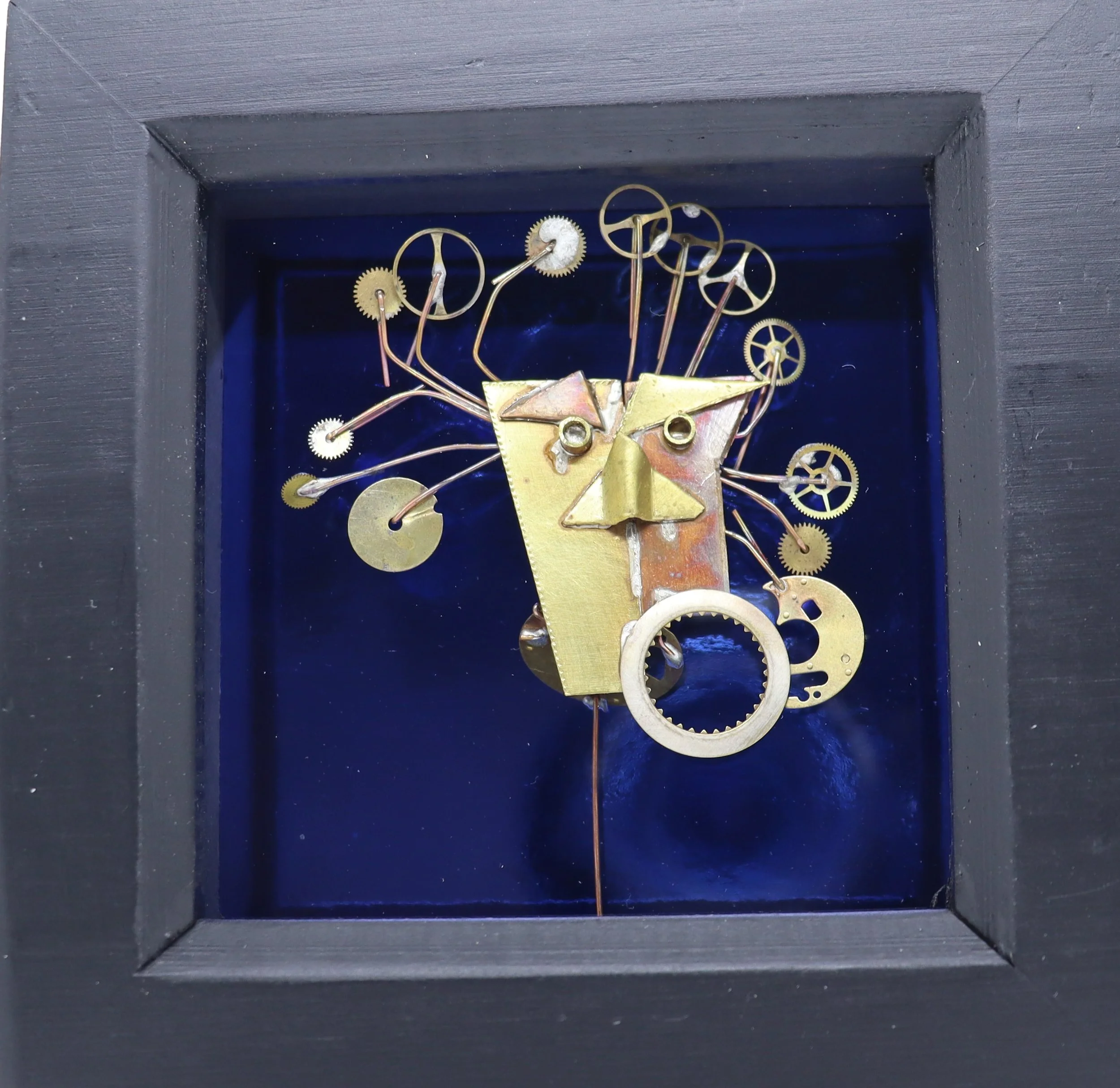 A clock sculpture with gears and metal parts forming a face, set inside a black frame.