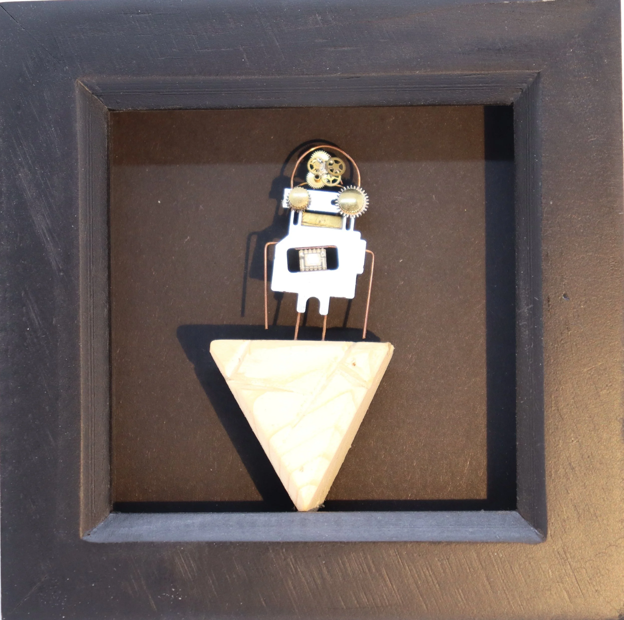 A small robot figure made of metal and electronic parts, standing on a triangular wooden base inside a black shadow box.