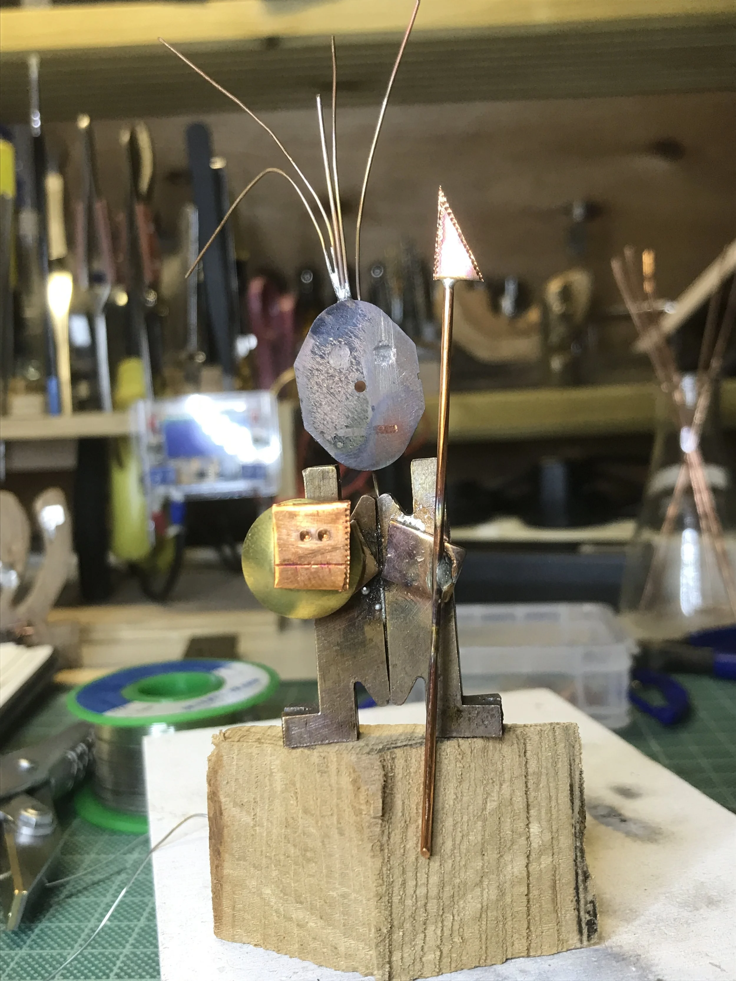 A small abstract sculpture made of metal parts, including wire, a small cube, and a triangular piece, mounted on a wooden block in a workshop setting.