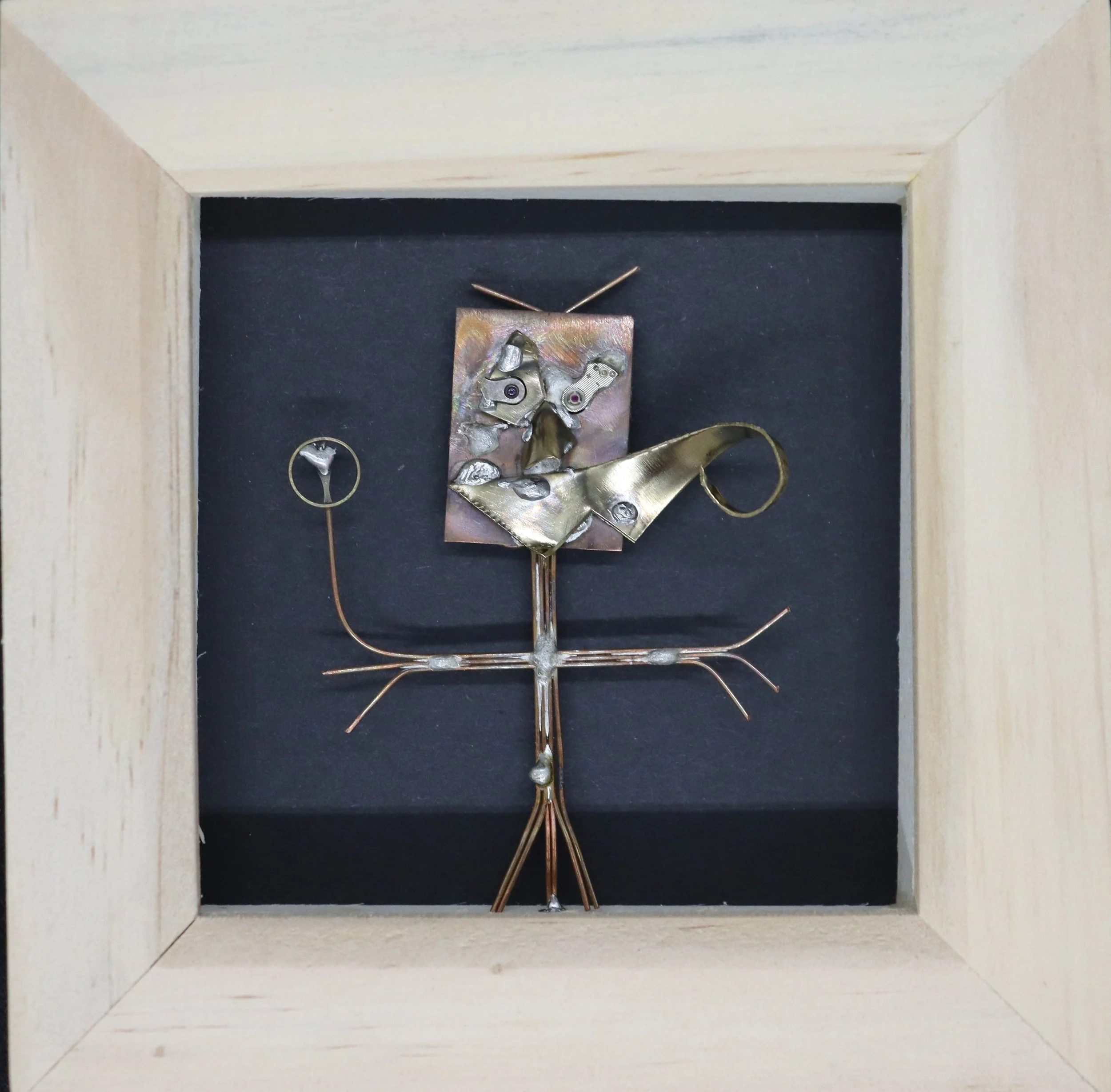 Metal abstract sculpture of a humanoid figure with a square head, circular eyes, and long limbs, displayed in a wooden frame with a black background.