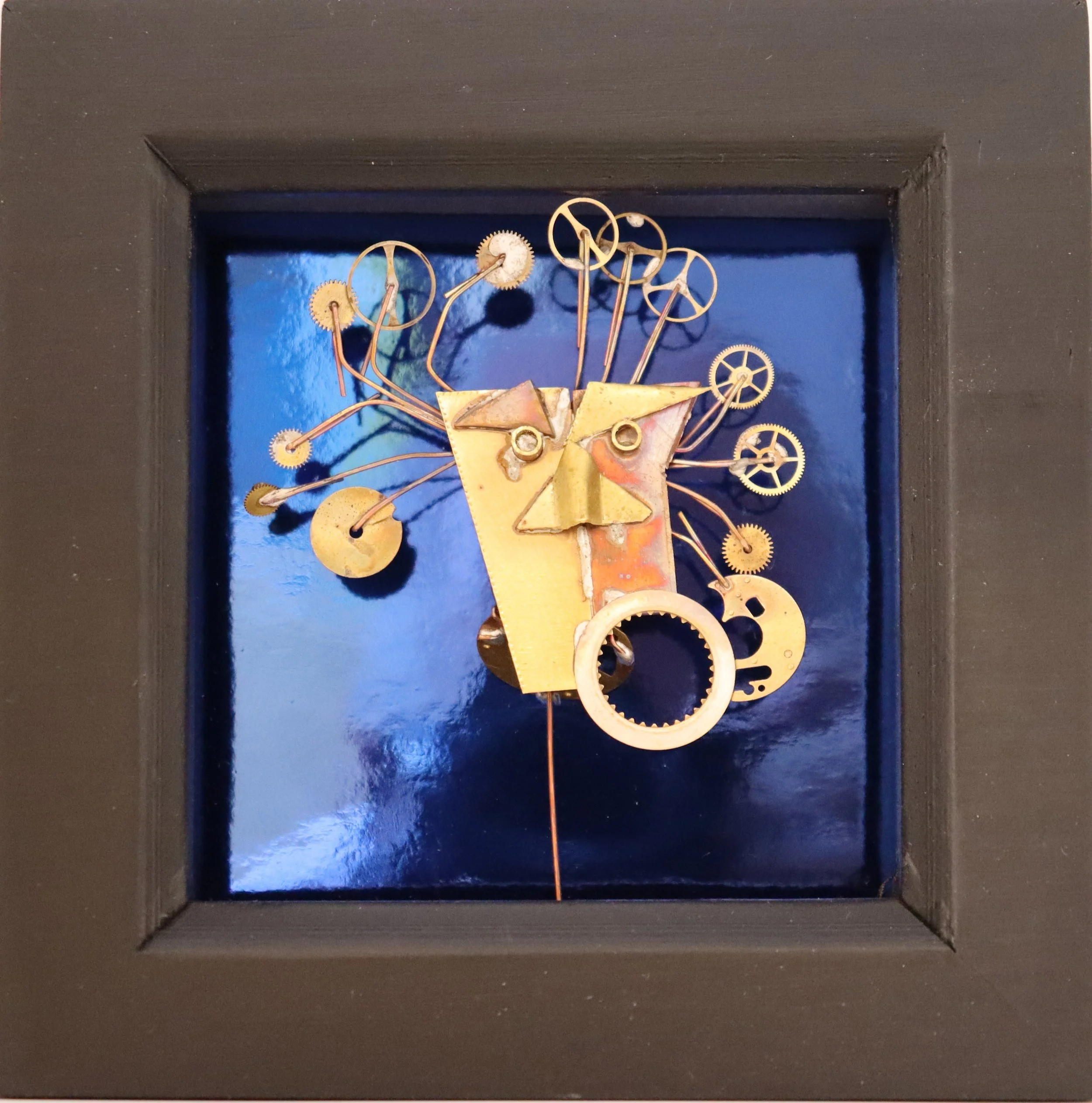 Mixed metal art piece resembling a face with gears and mechanical parts, framed in a black square frame with a reflective blue background.