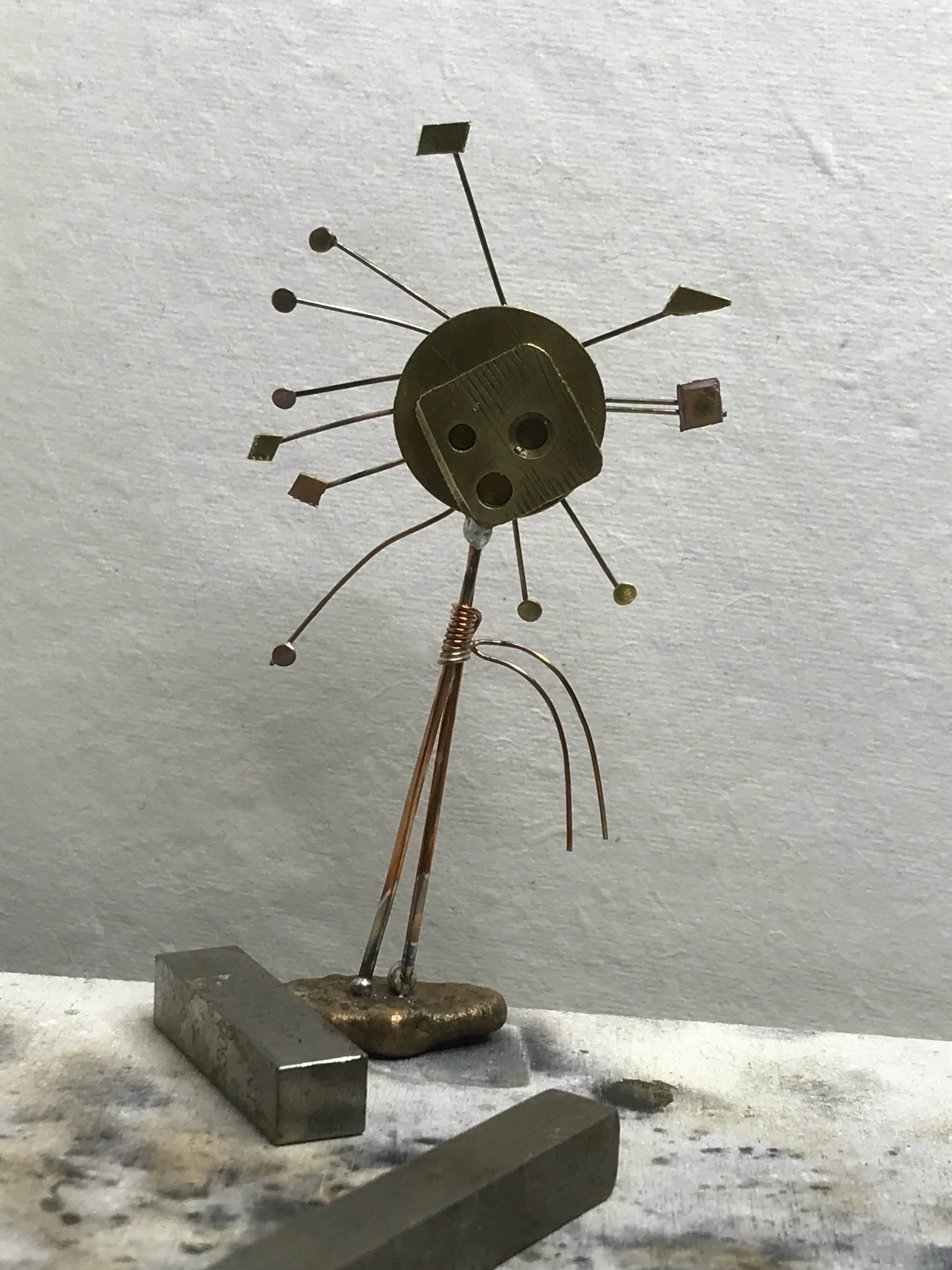 A small handmade sculpture of a person made with various metal parts, including a round face with holes for eyes and mouth, arms, legs, and a base, standing on a surface with metal pieces around it.