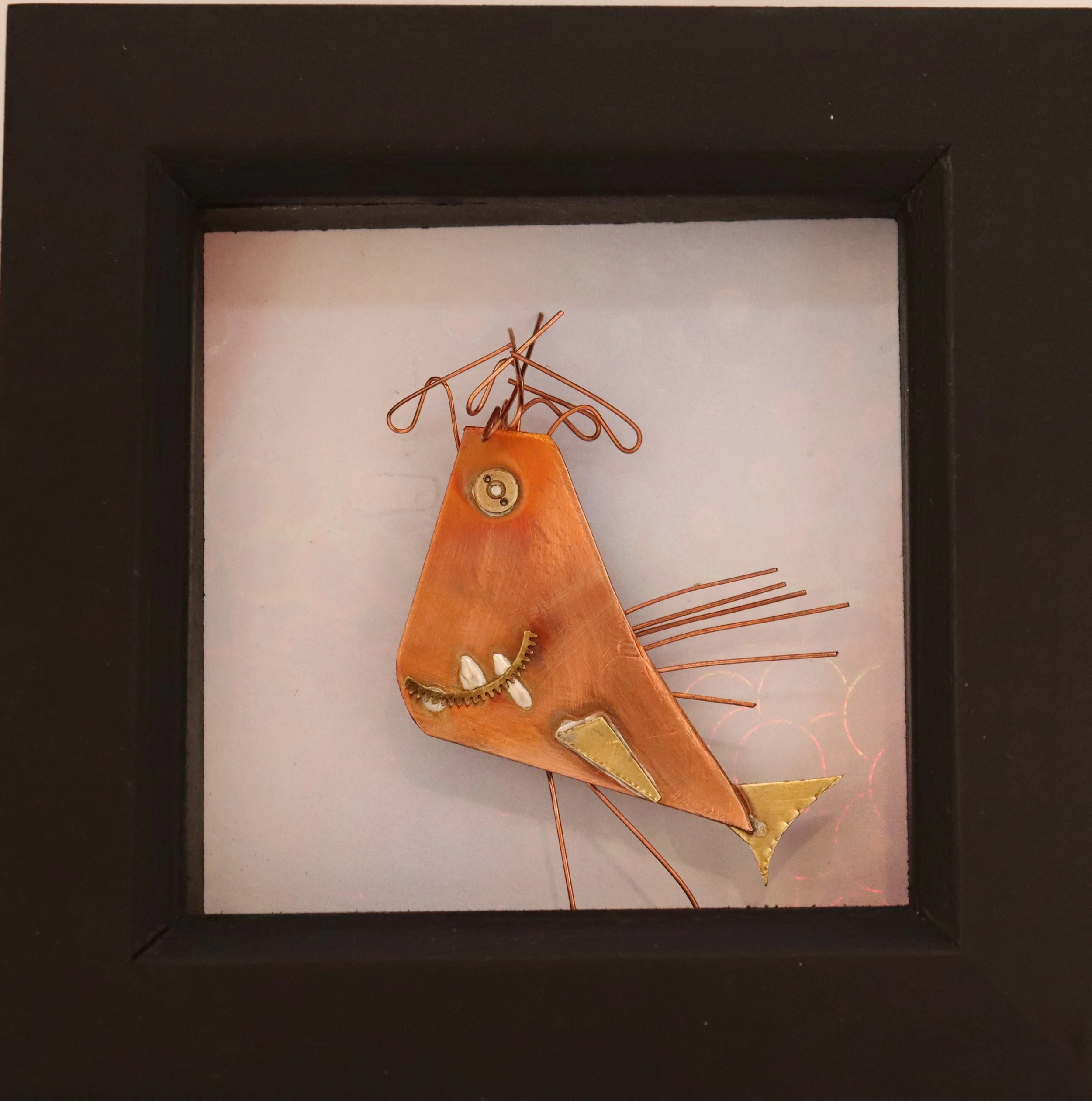 Folk art sculpture of a fish made from copper, metal, and other materials, displayed in a shadow box frame.