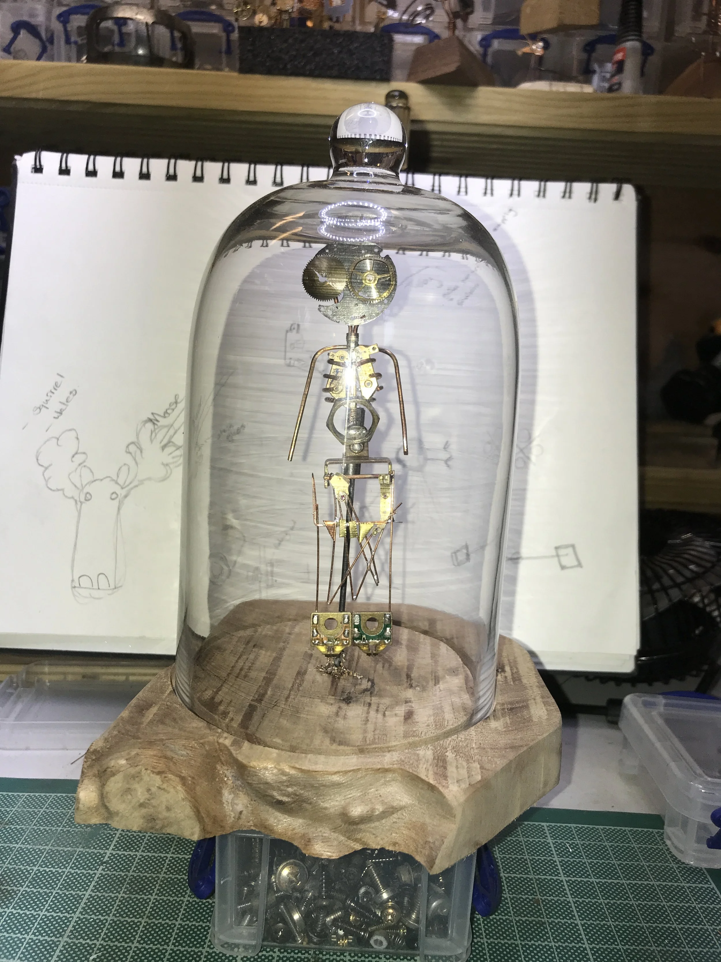 A vintage vacuum tube with metal components inside, encased in a glass dome, placed on a wooden base in a workshop.