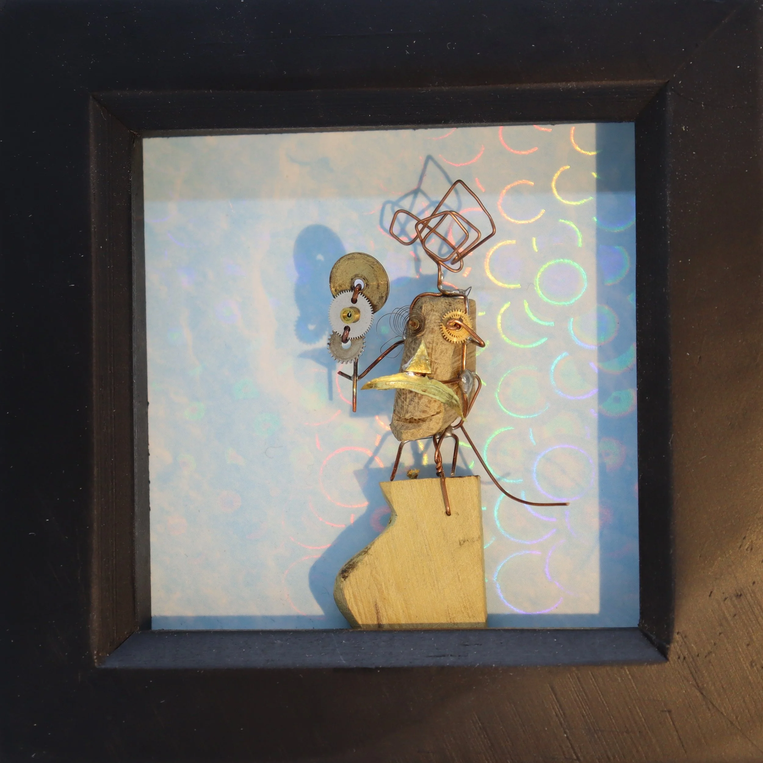 Mixed media art piece of a bird sculpture made from metal and wood, holding a paint palette, inside a shadow box with holographic circles reflected on the background.