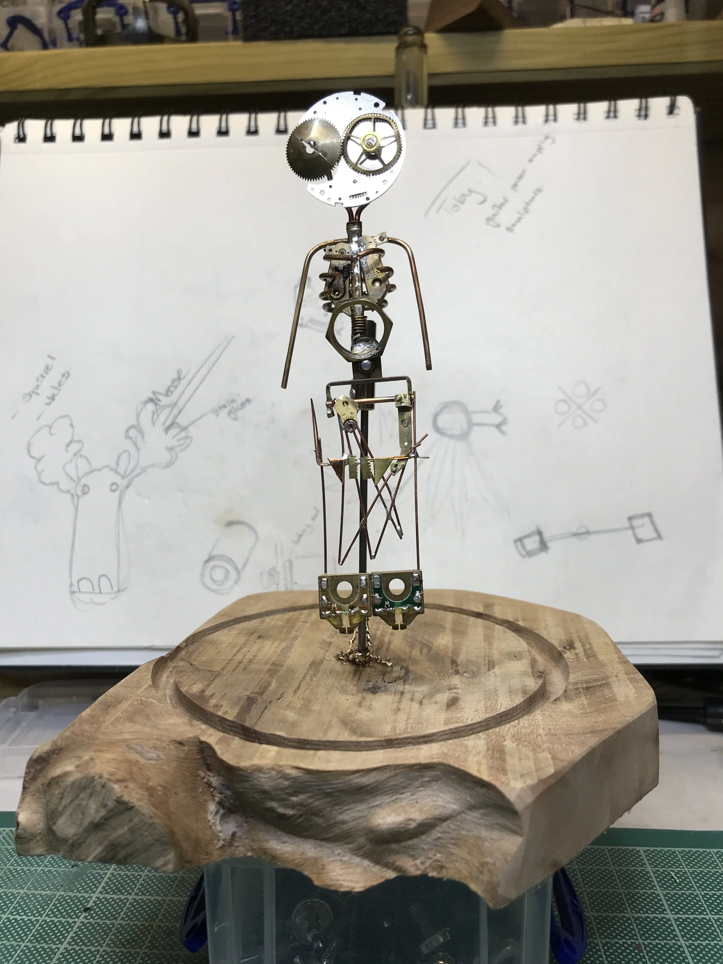 A humanoid sculpture made from various metal watch parts and gears, standing on a wooden base.