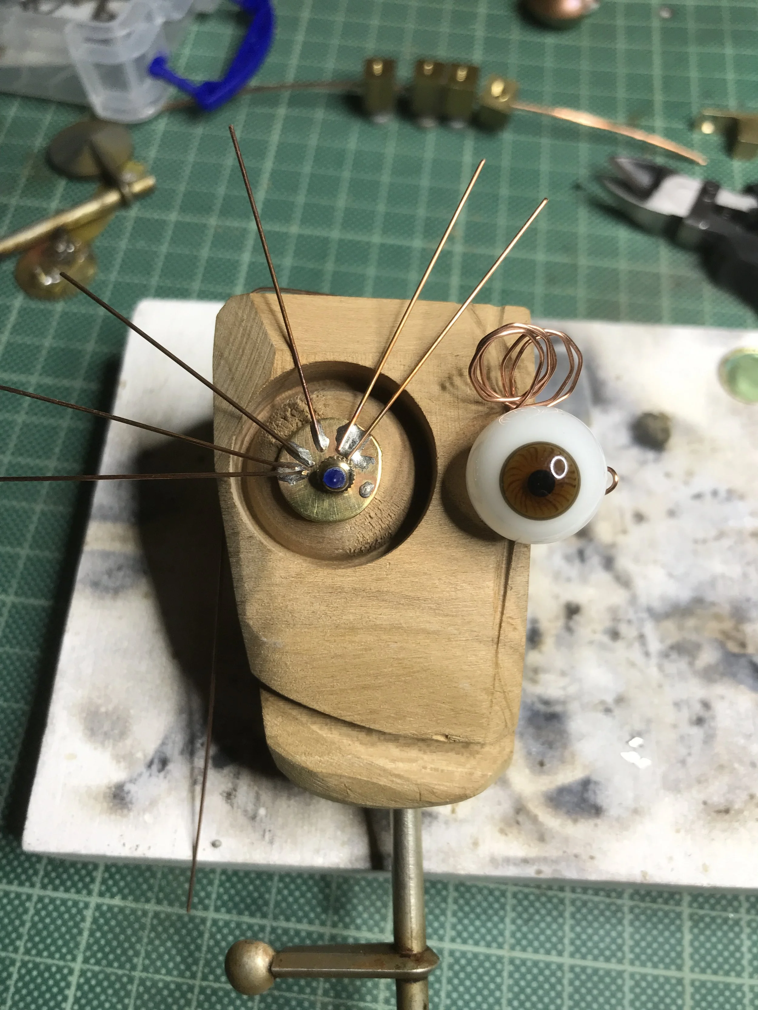 A handmade eye sculpture with brown and white glass eye, copper wire coil, mounted on wooden block, surrounded by various metal tools and parts.