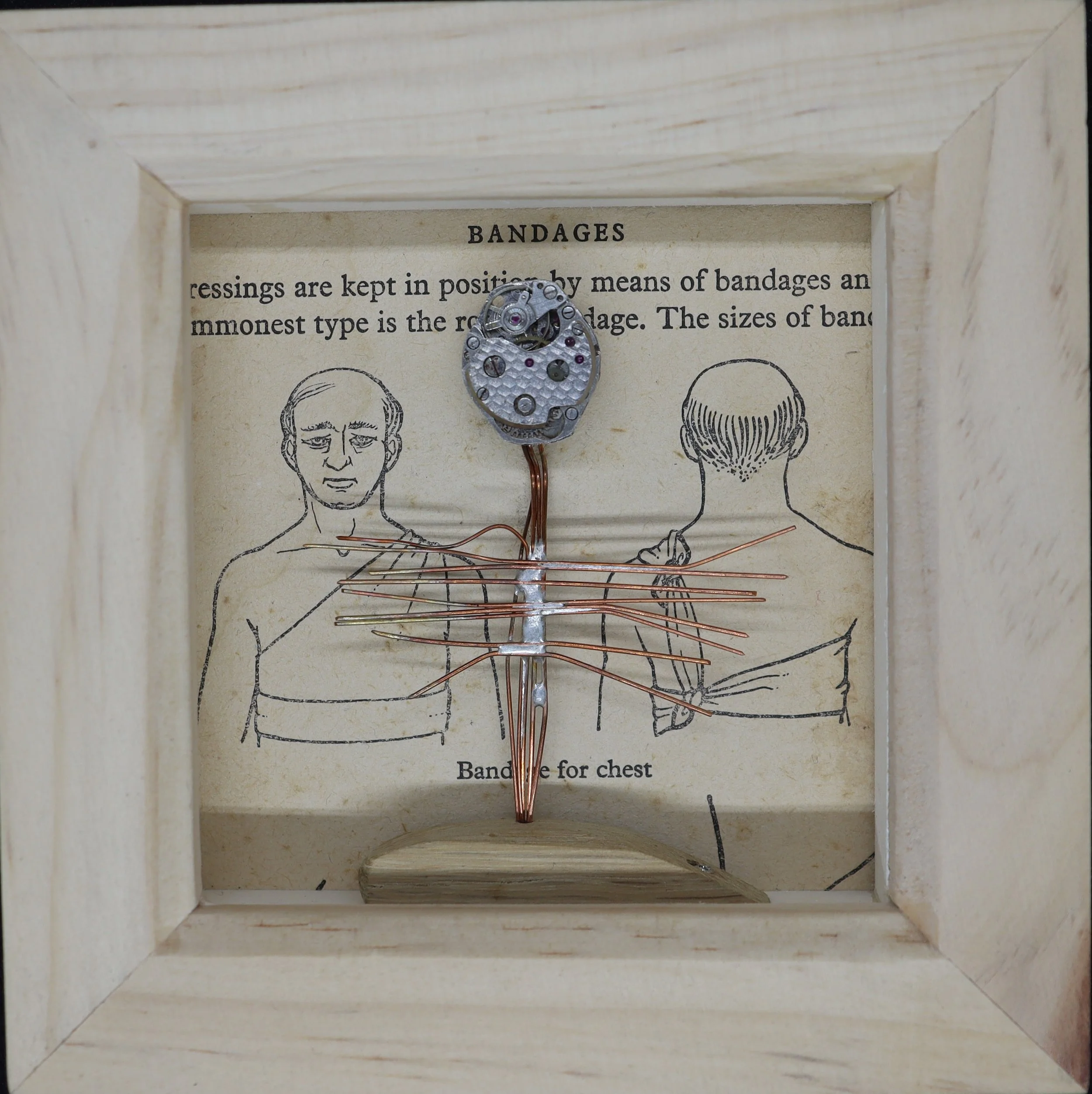 A small mechanical watch movement mounted on a wire stand inside a wooden display box. Behind it is a vintage diagram of a human chest showing placement of bandages for dressing wounds.