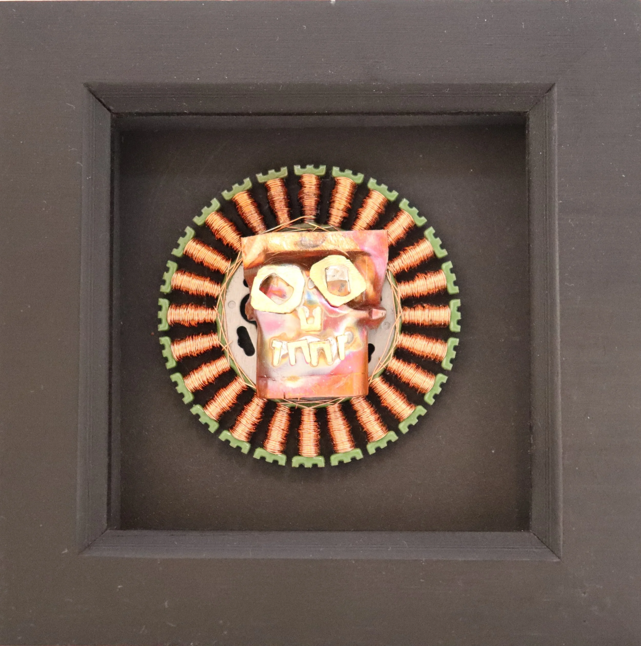 Mixed media art piece resembling a skull with metallic components, placed on a circular base with radiating copper wire coils, displayed in a black shadow box frame.