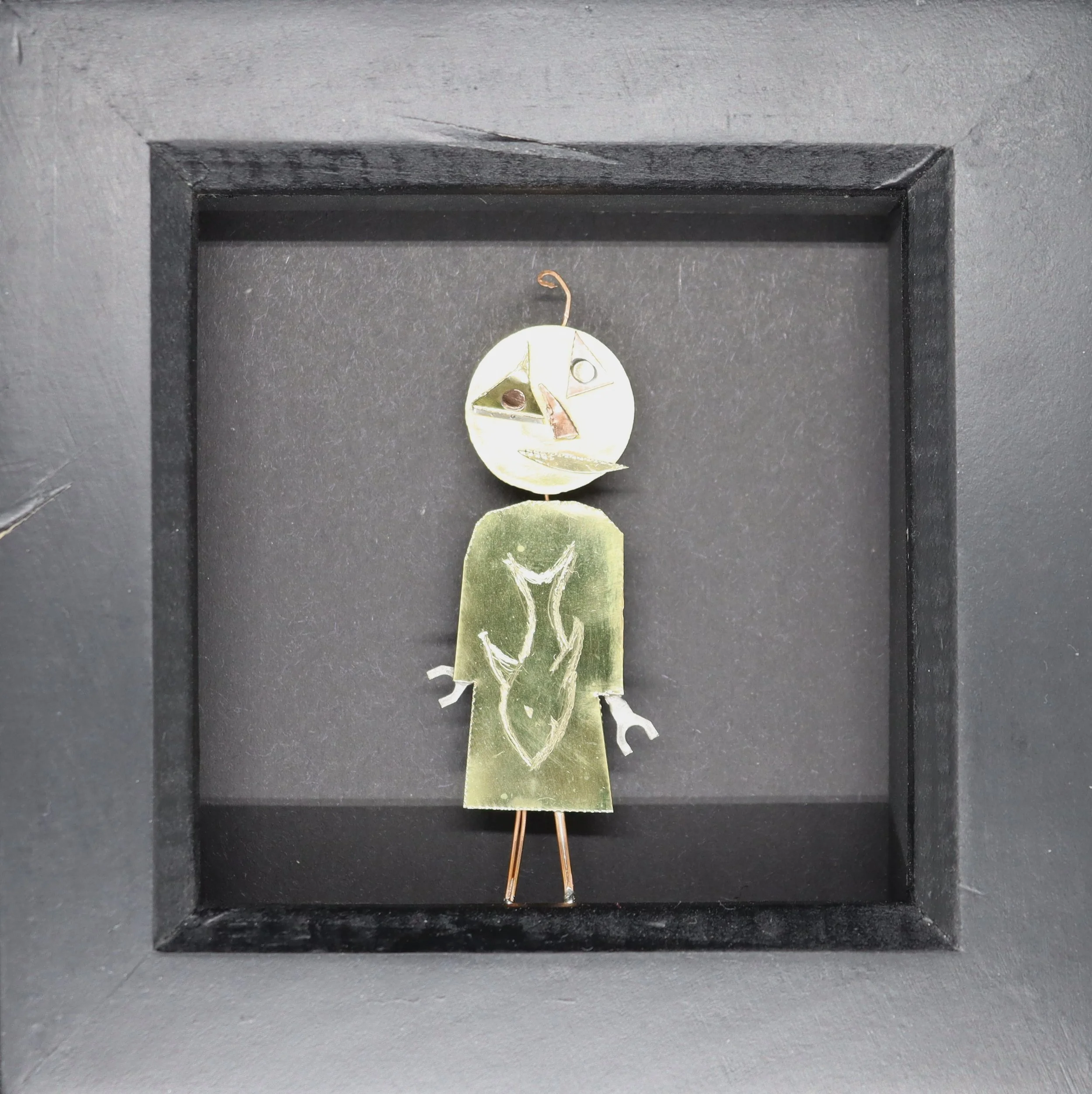 A small art sculpture resembling a humanoid figure with a circular head and a green body, displayed in a black shadow box. The face is stylized with geometric shapes for eyes, nose, and mouth.