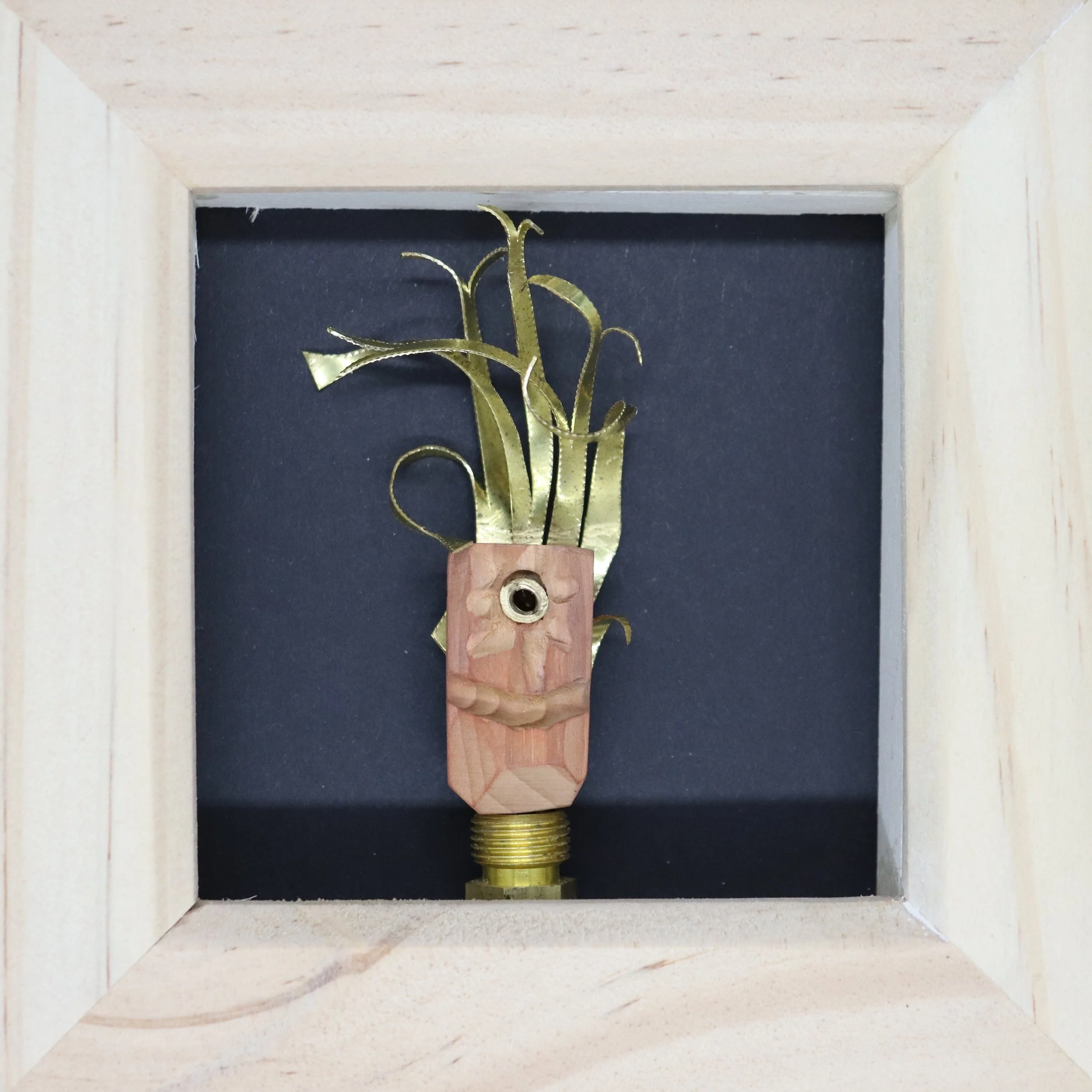 A framed artwork featuring a light-colored wooden block with a smiling face carved into it, and metal elements resembling plants and leaves growing from the top, set against a dark background.