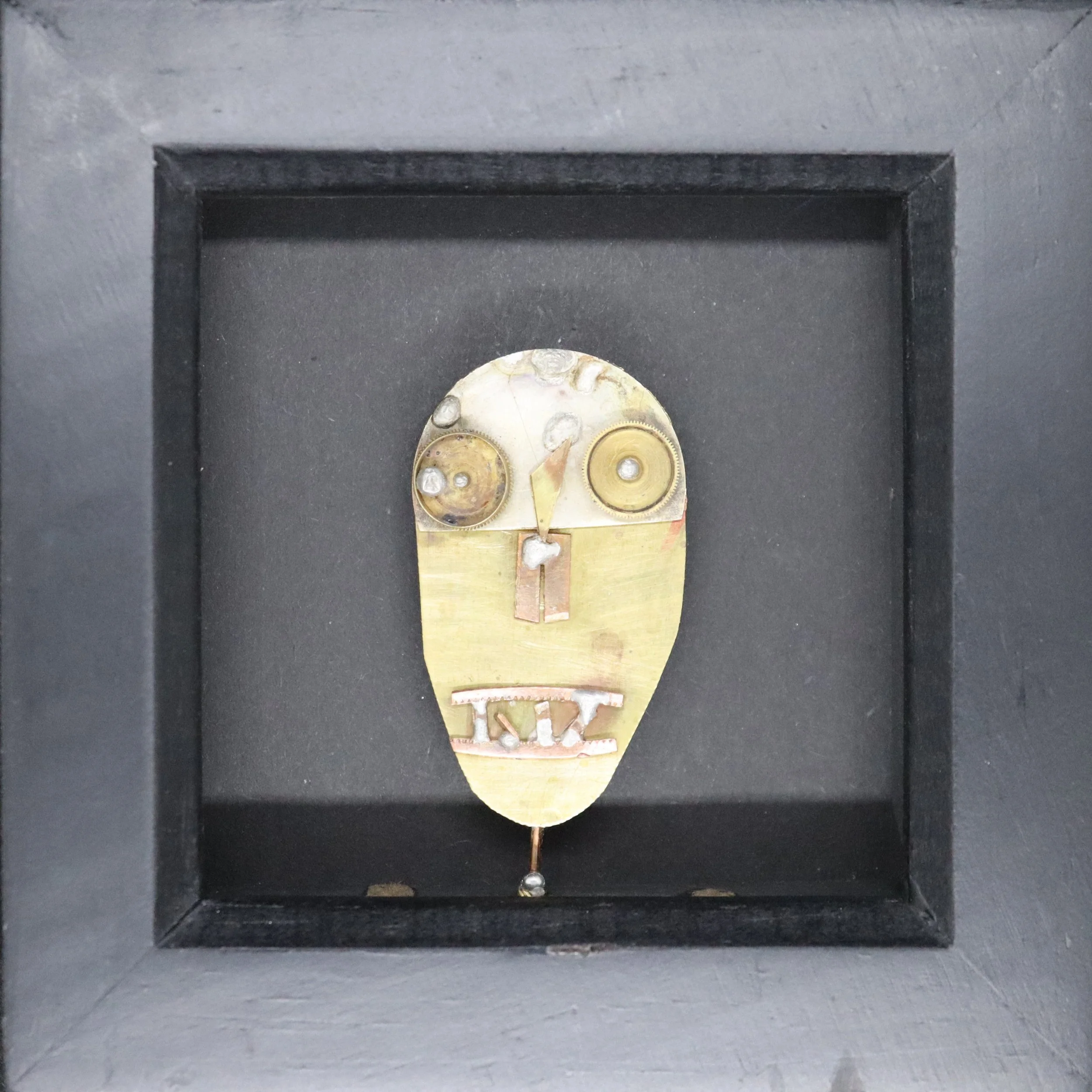 A mixed media sculpture of an abstract face with gear eyes, on a black background.