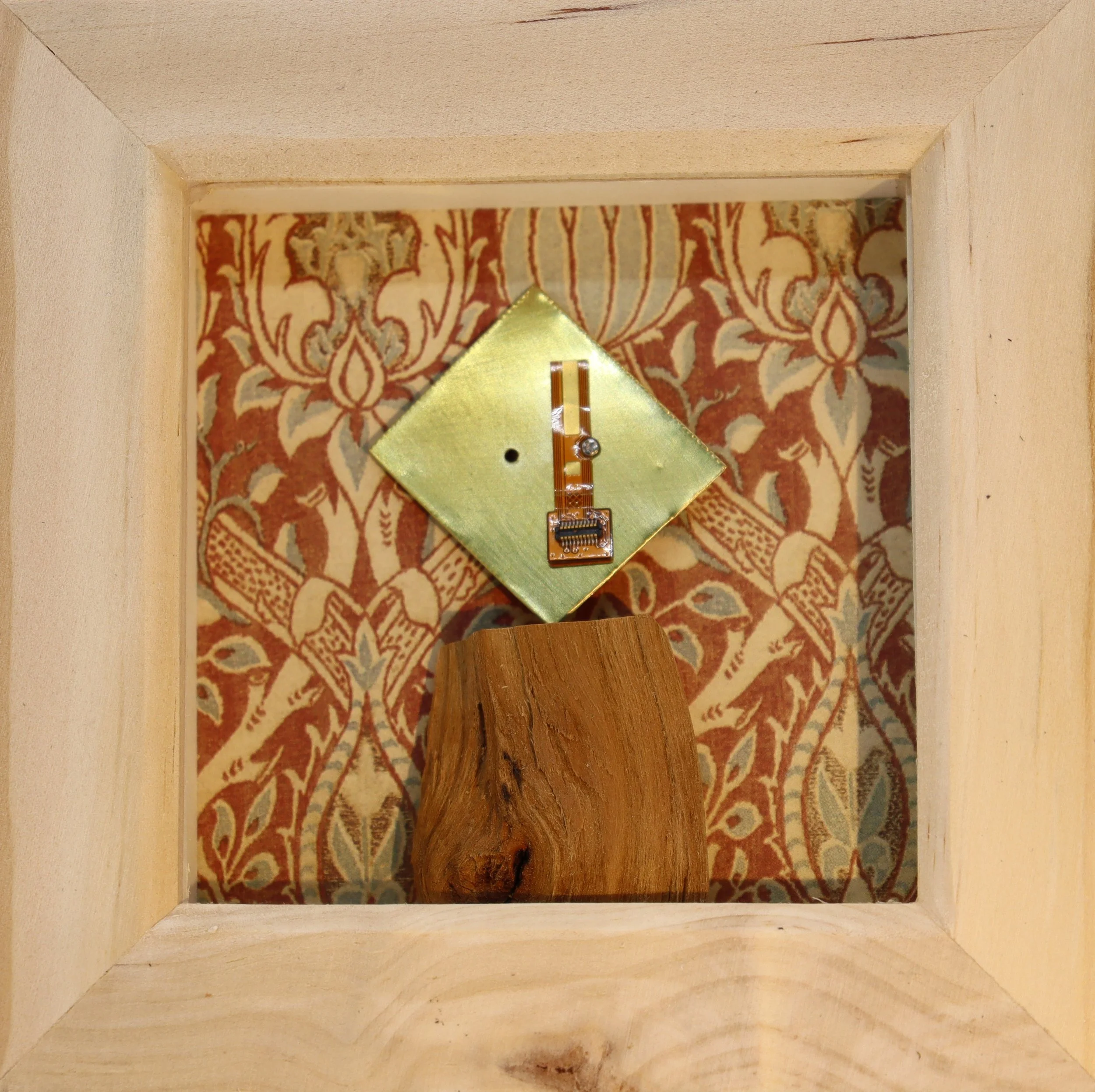 Inside a wooden shadow box, there is a fabric-lined background with a floral pattern. A small electronic component with a gold square base and a ribbon cable is mounted on the fabric, and a piece of wood is placed at the bottom.