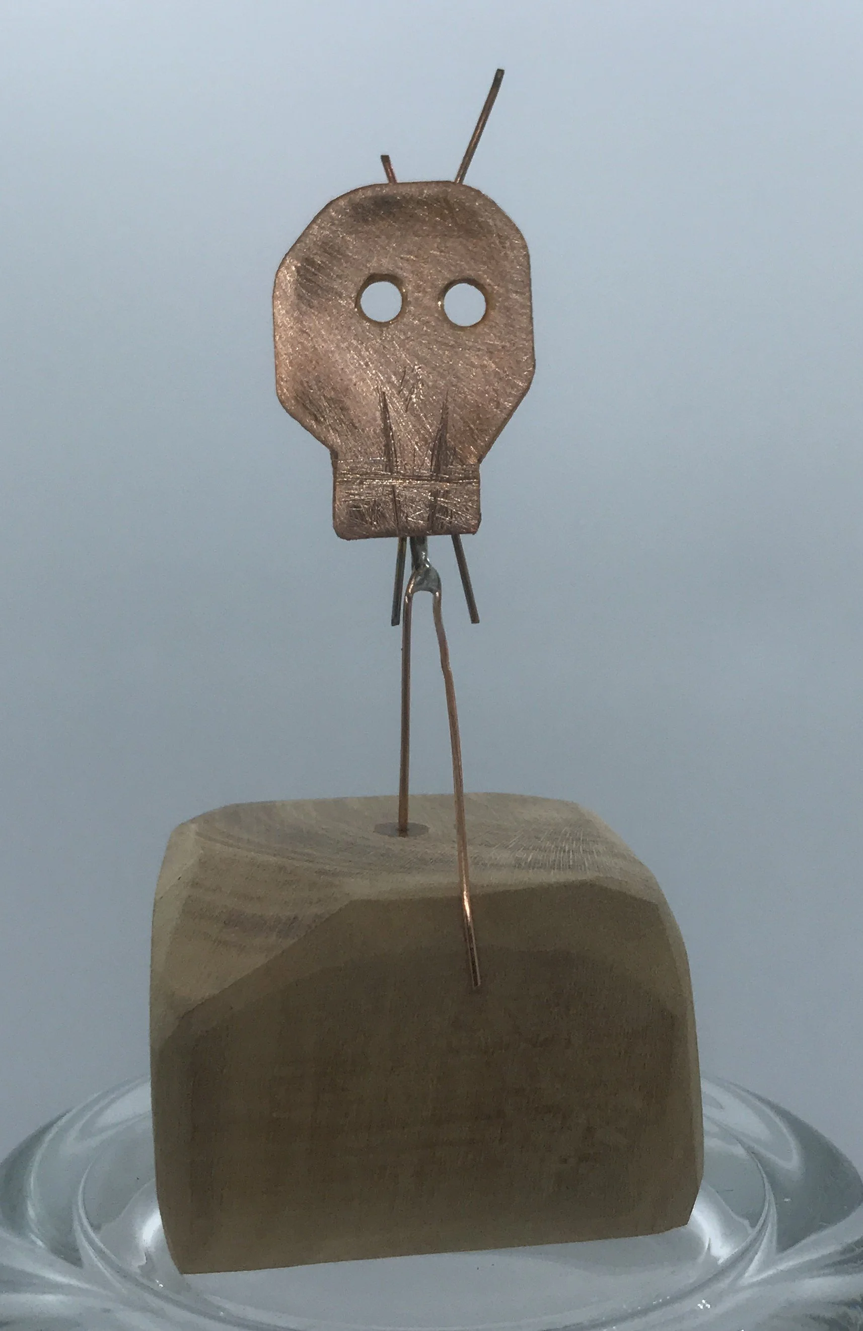A small skull-shaped object made of metal, with two circular holes for eyes and carved lines resembling teeth, mounted on thin metal wires attached to a wooden block, displayed on a glass surface.