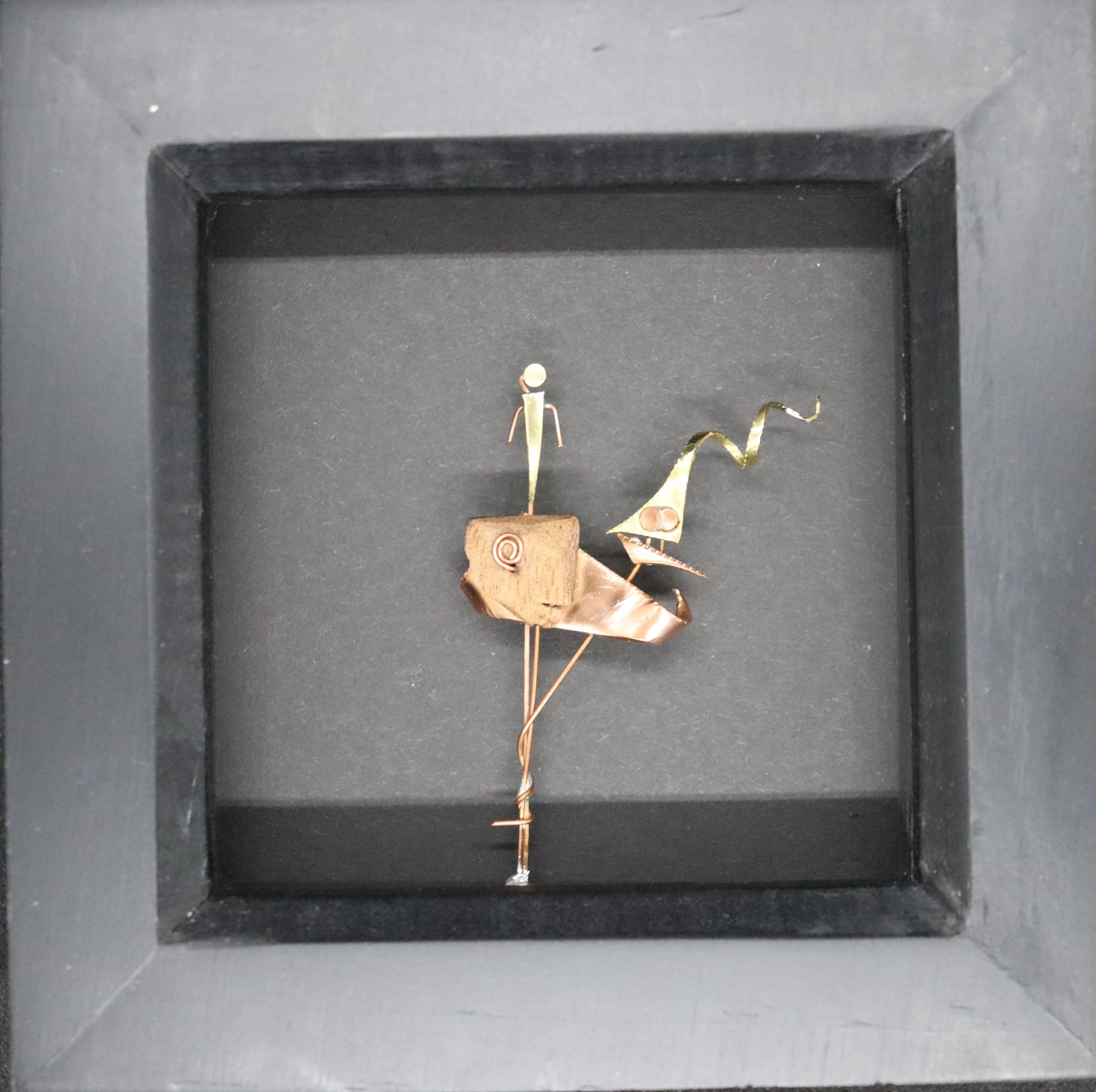 Small art sculpture of a person sitting on a stylized animal with a curved tail, made of mixed materials including wood and metal, displayed inside a black shadow box.