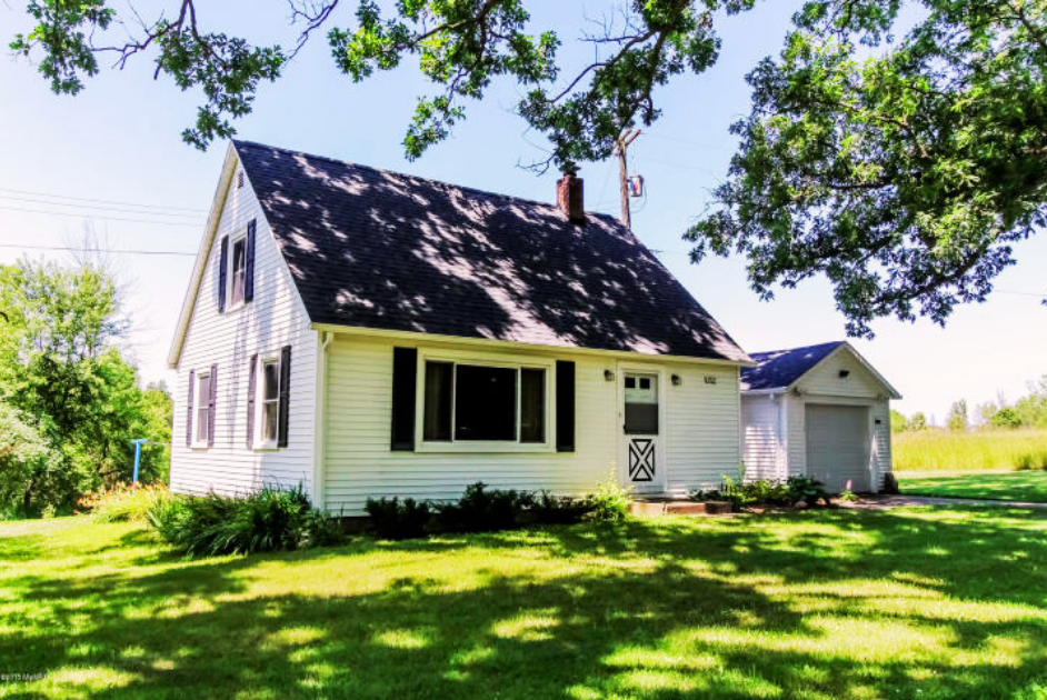 A white house with black shutters and a dark roof, surrounded by green grass and trees, with a detached garage in the background on a sunny day.