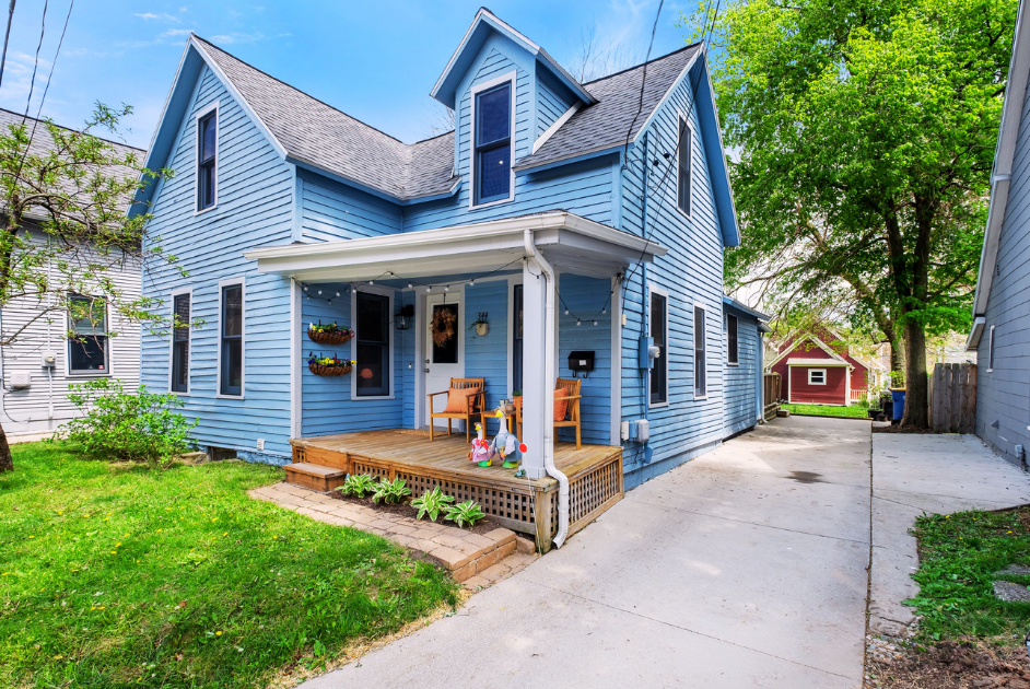 Blue two-story house with a front porch, two wooden chairs, decorative string lights, and small potted plants, surrounded by green trees and grass.