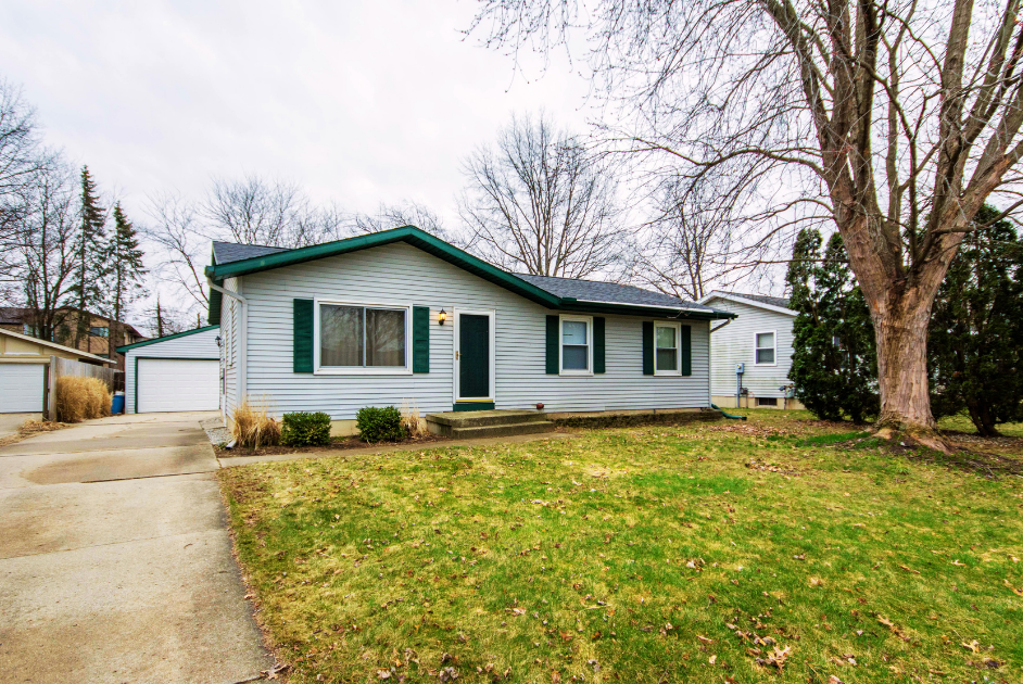 A light blue single-story house with green shutters, a front porch with steps, a driveway leading to a garage, and a large tree in the front yard. The house is situated in a suburban neighborhood with other houses and trees around.