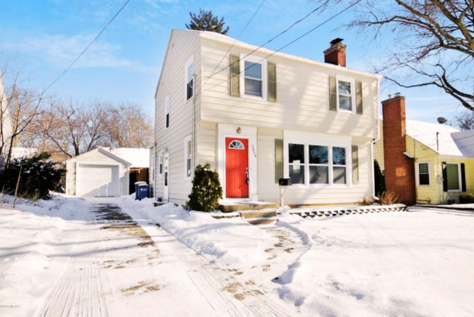 A white, two-story house with a red front door in a snowy neighborhood, with a detached garage and neighboring houses visible, clear blue sky.