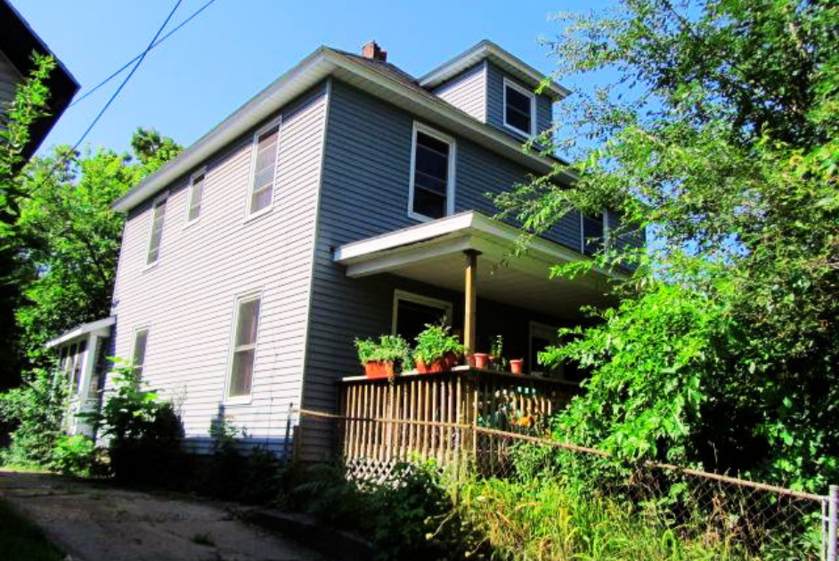 A two-story house with gray siding and multiple windows, surrounded by green trees and shrubs, featuring a small wooden porch with potted plants.