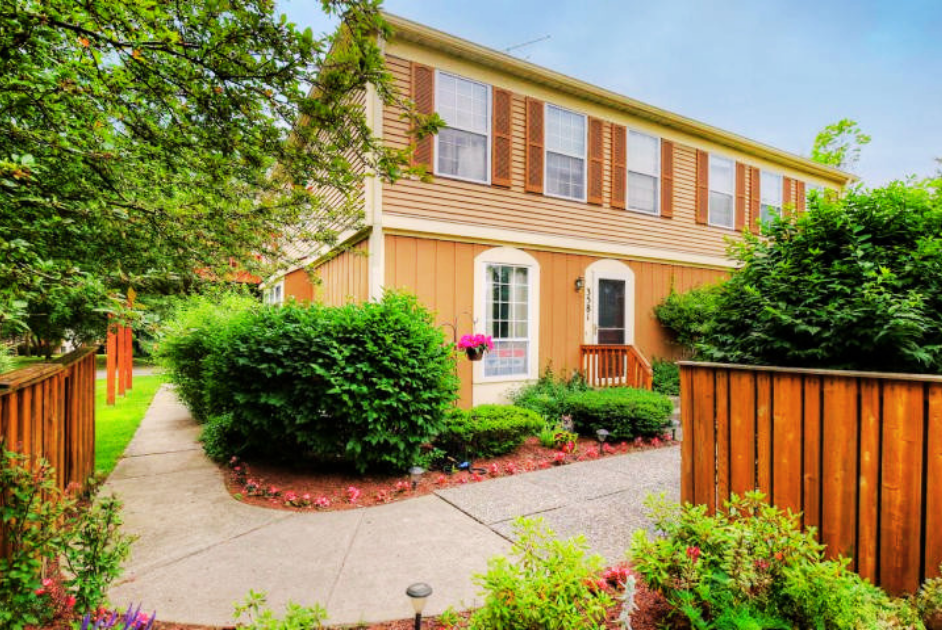 Front yard of a two-story house with beige siding, white trim, and brown shutters, surrounded by green bushes and colorful flowers, with a concrete walkway and wooden fences on either side.