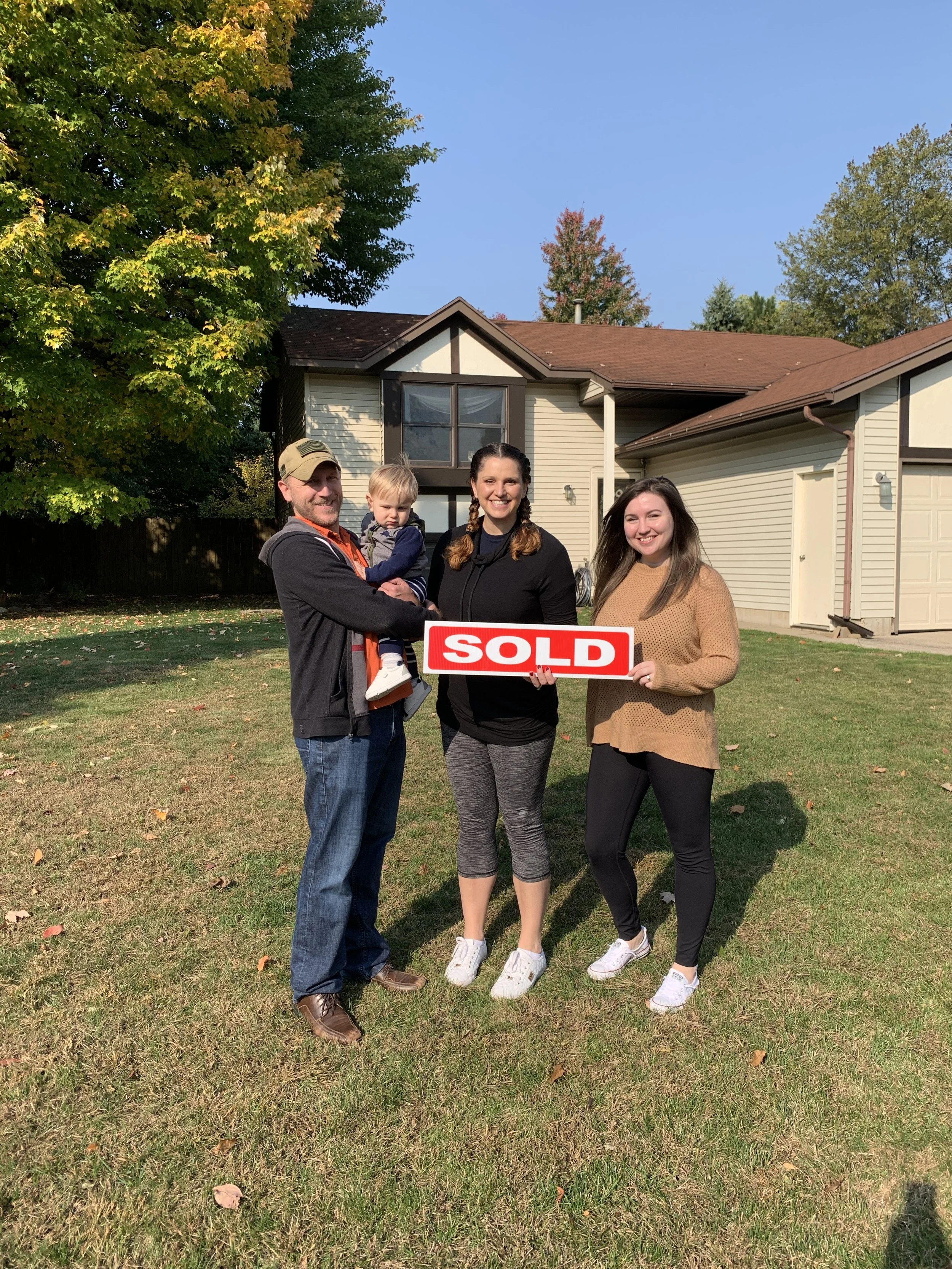 A family of four standing outside in front of a house. The man on the left is holding a young boy, both smiling. The woman in the middle is smiling, and the woman on the right is holding a