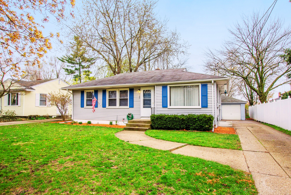 A suburban house with a manicured lawn and a curved concrete walkway leading to the front steps. The house has blue window shutters, a small porch, and a white picket fence on the right side. Trees with mostly bare branches surround the house, indicating fall or early winter.