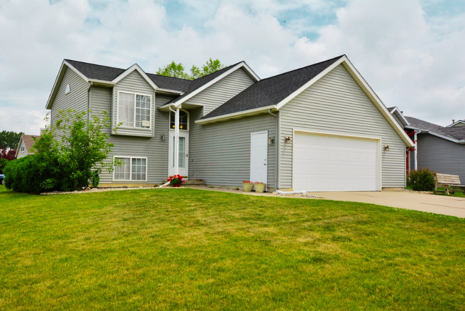 A two-story suburban house with gray siding and a white garage door, surrounded by a well-maintained front lawn and some bushes, under a partly cloudy sky.