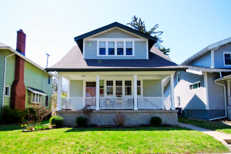 Front view of a white two-story house with a covered porch, railing, and multiple windows, flanked by neighboring houses and a grassy yard.