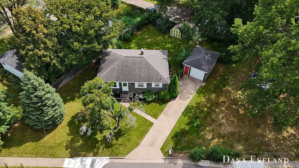Aerial view of a house with a grey roof and white exterior, surrounded by green trees and a lawn. There is a concrete driveway leading to a detached garage with a red door. A small garden shed is visible behind the garage. The front yard has a large tree near the sidewalk and a sidewalk running along the street.