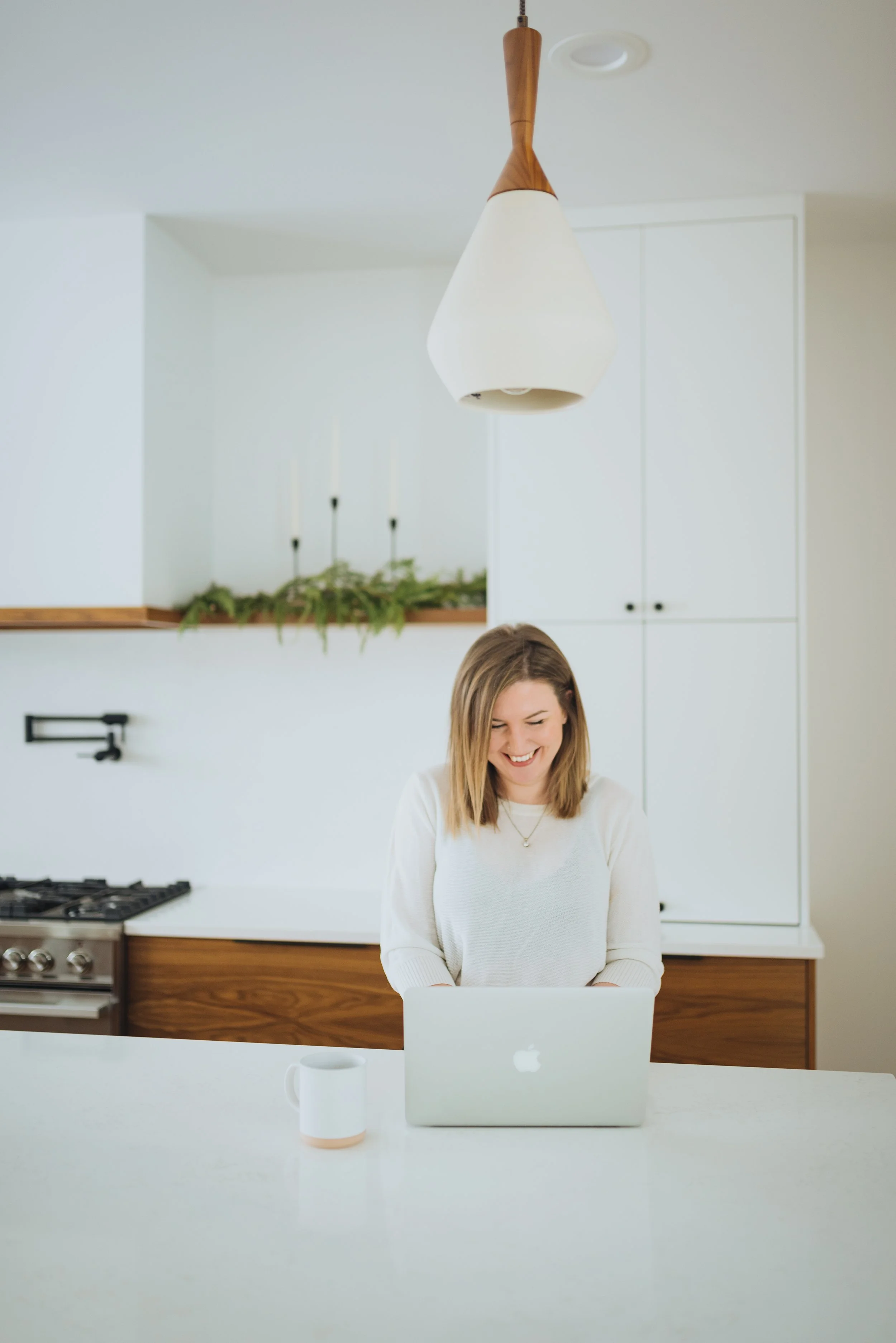 Woman smiling and working on a laptop in a modern kitchen with white cabinets and a wooden countertop.