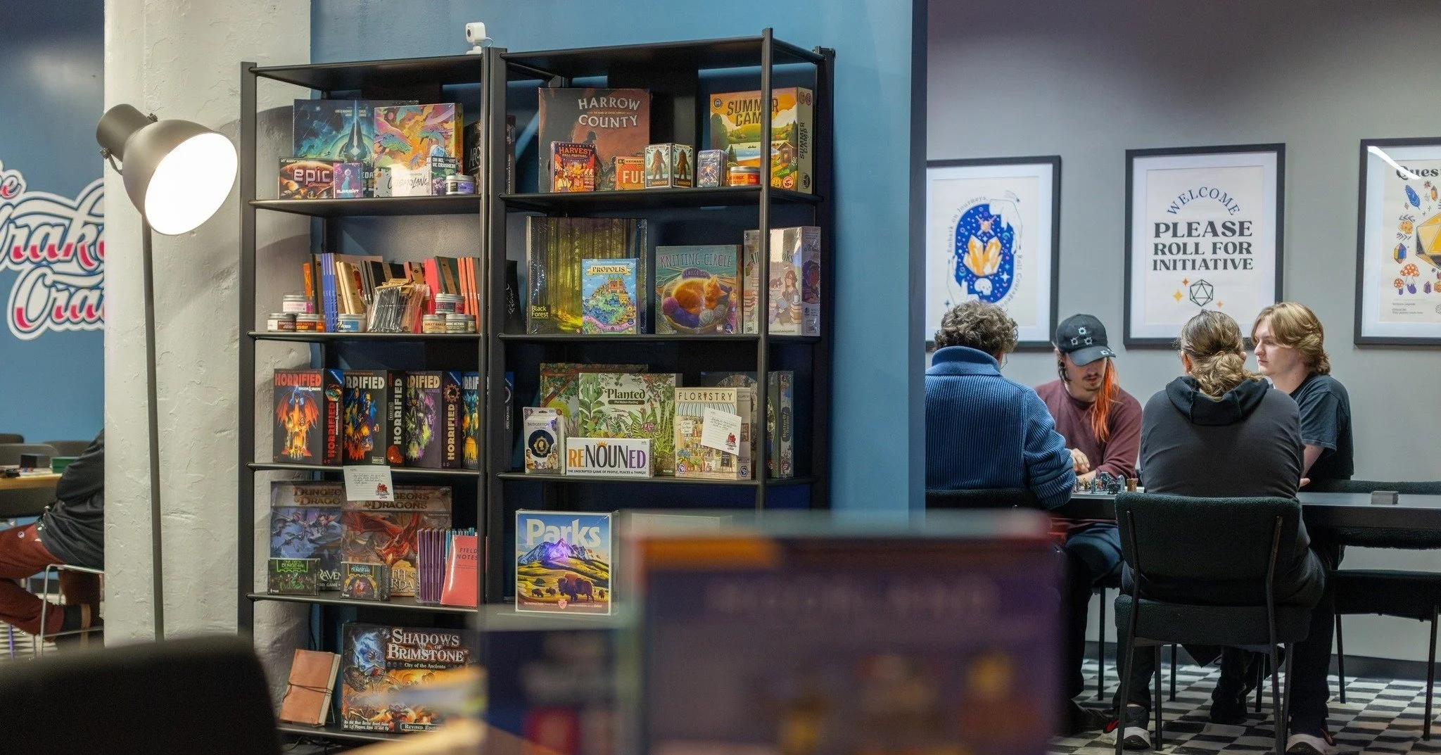 A bookshelf filled with board games and books in a gaming store, with customers playing a game at a table behind it, and framed posters on the wall encouraging a gaming initiative.