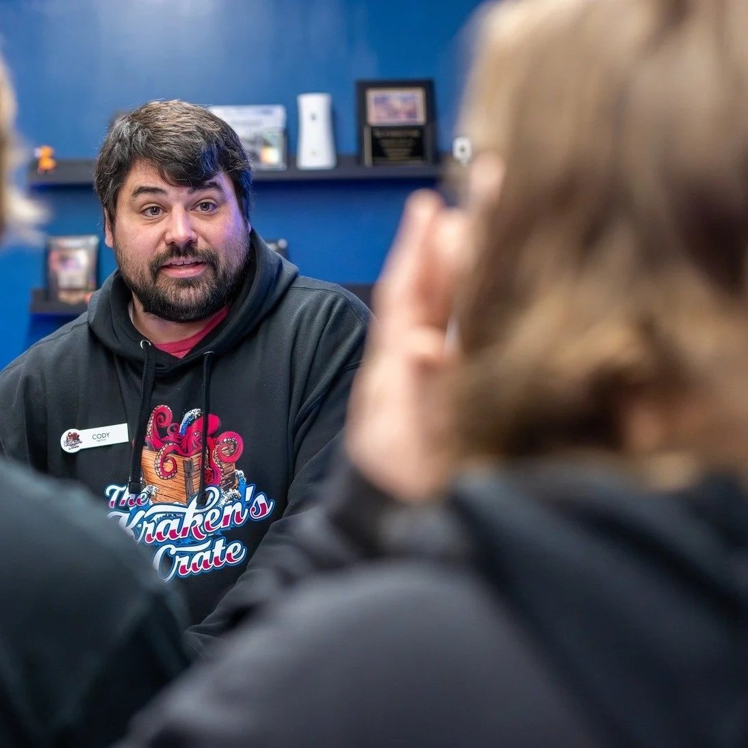 A man with a beard and dark hair wearing a hoodie with a embroidered logo, speaking to a group of people in an indoor setting.