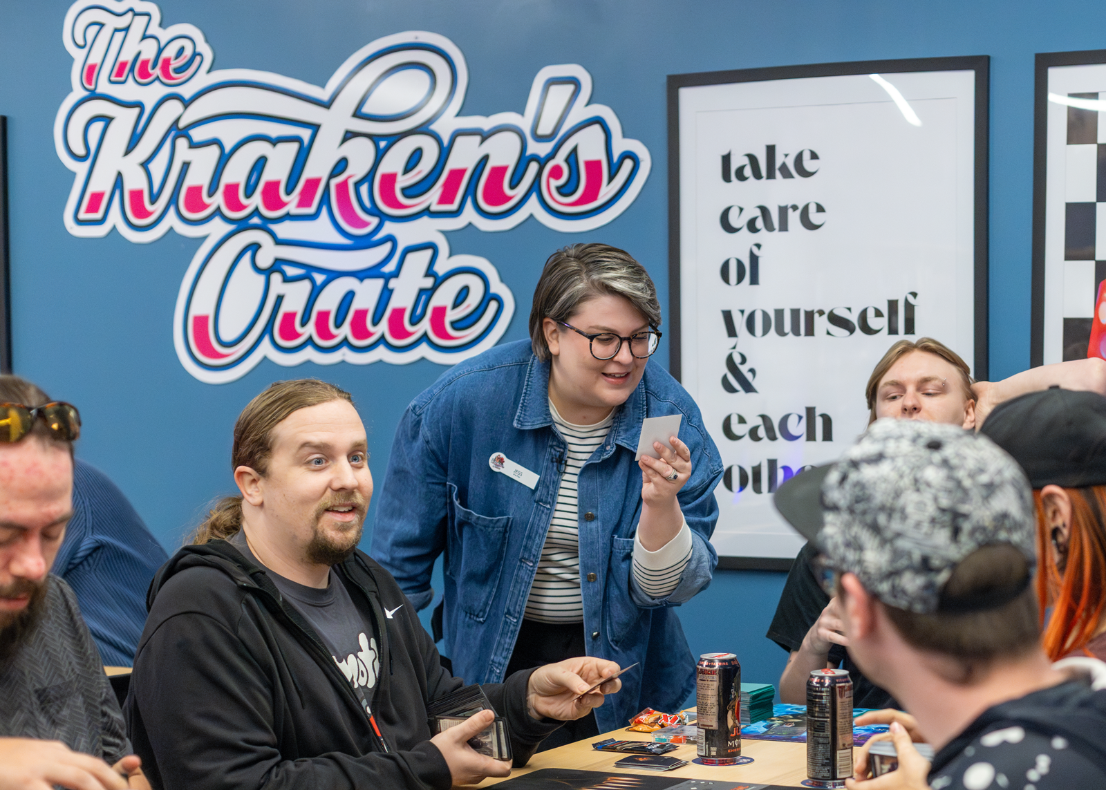 Group of people sitting around a table playing a card game in a room decorated with signs that read 'The Kraken's Crate' and 'take care of yourself & each other'.