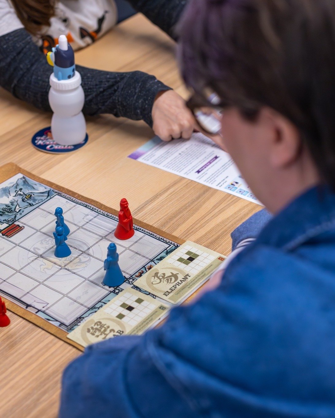 A person playing a board game involving blue and red pieces, with a game sheet labeled 'Elephant,' on a wooden table.