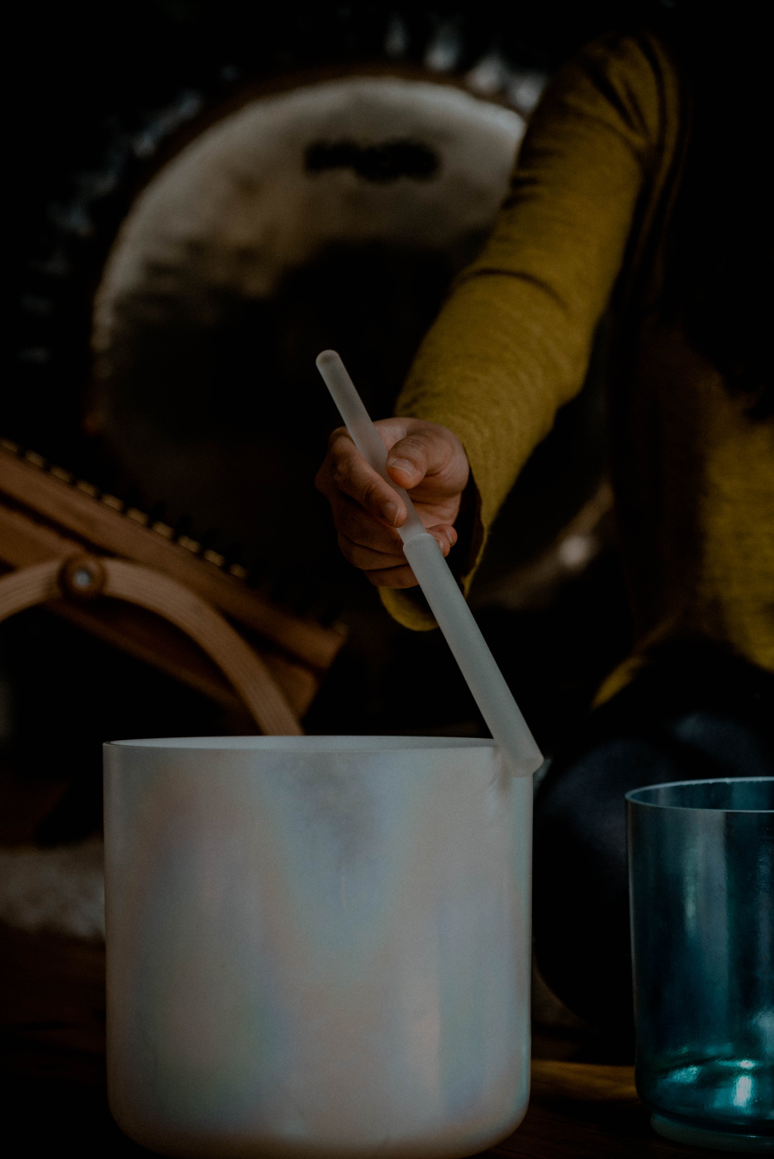 A person wearing a yellow sweater and striped pants is holding a white straw and putting it into a white mug filled with a beverage. There is a blue glass to the right of the mug, and a guitar is visible in the background.