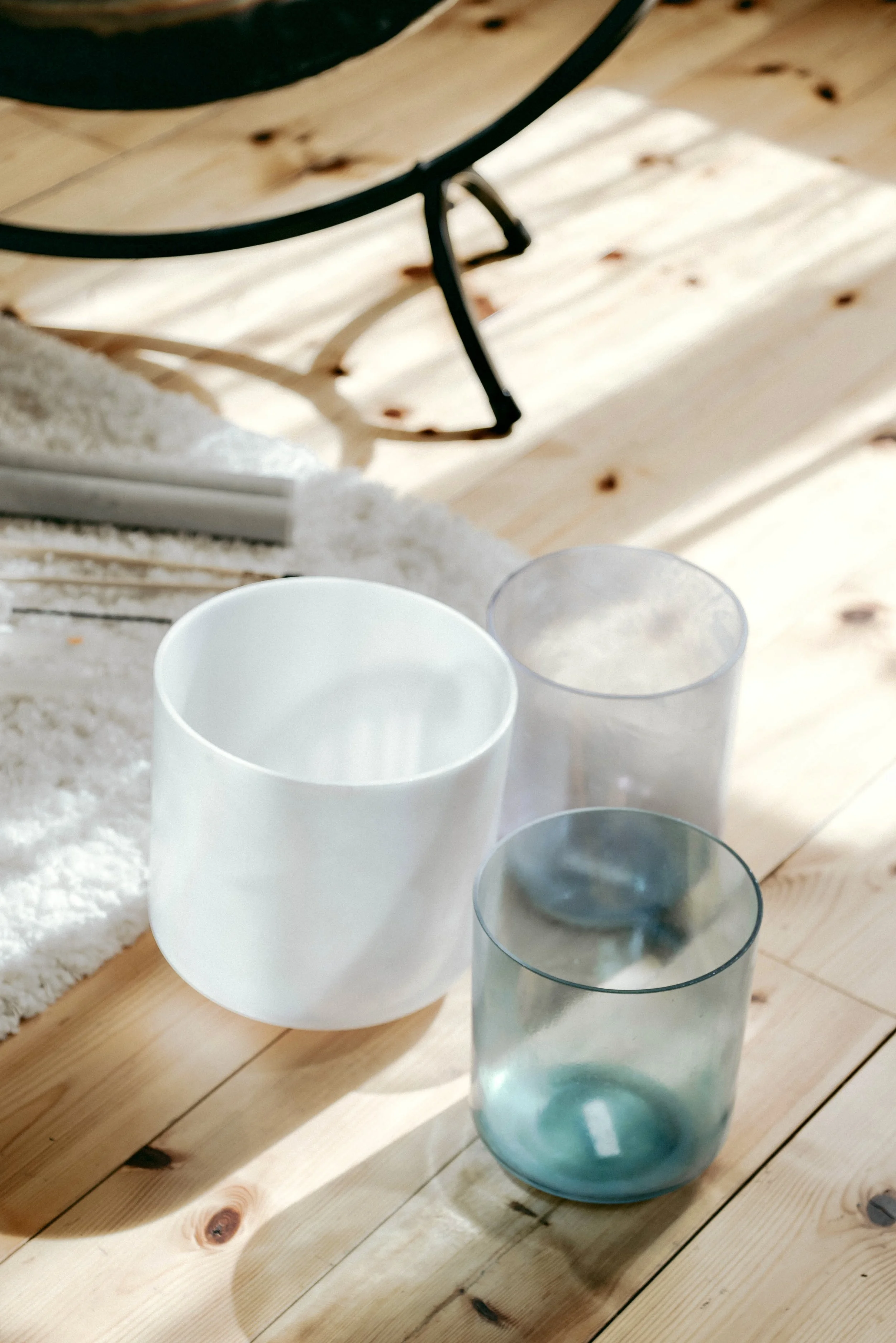 Four glasses on a wooden table, with two appearing to contain water and the other two empty. In the background, a black metal object casting shadows on the light-colored wood surface.