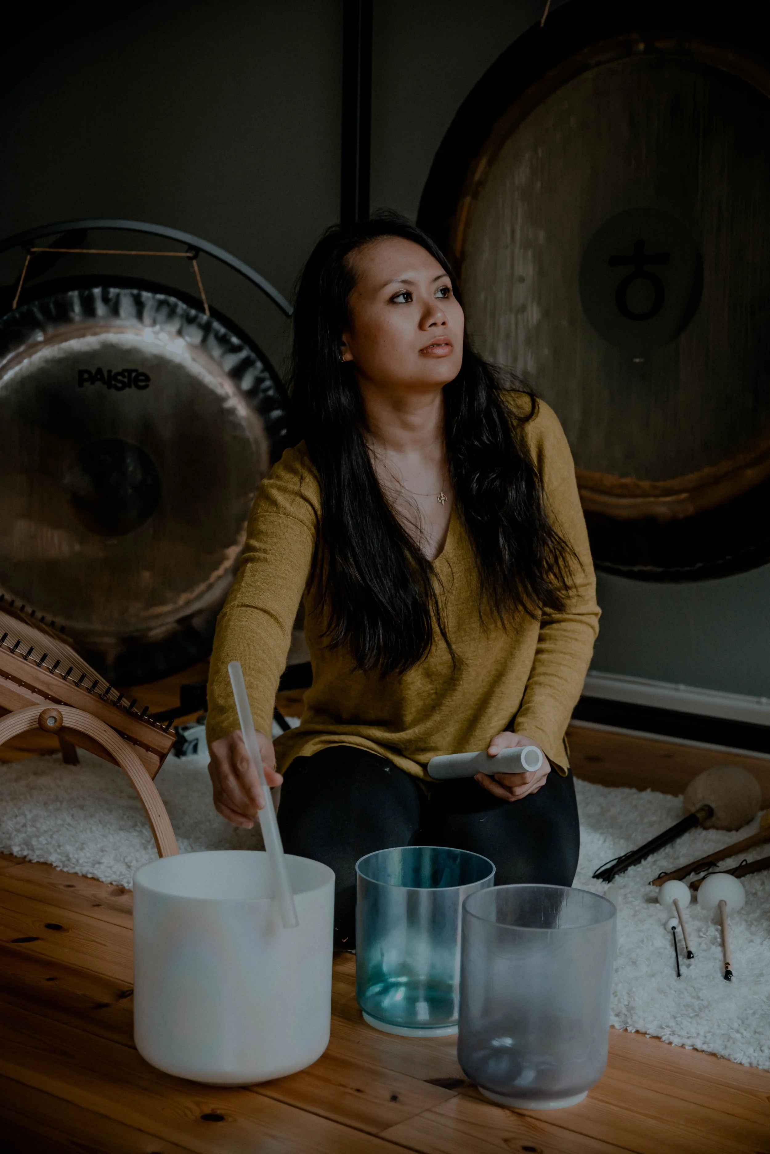 A woman with long dark hair sitting on the floor, surrounded by musical instruments and glass cups, with a large gong and a drum in the background.