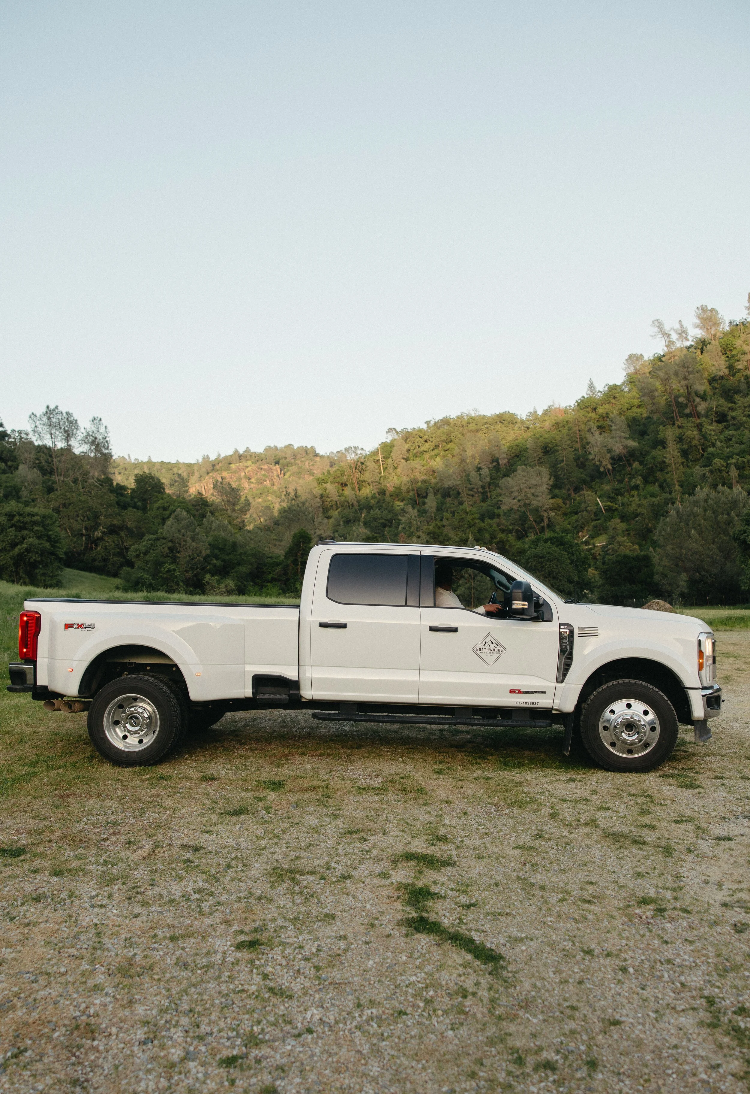 A white pickup truck parked on a gravel surface with greenery and hills in the background.