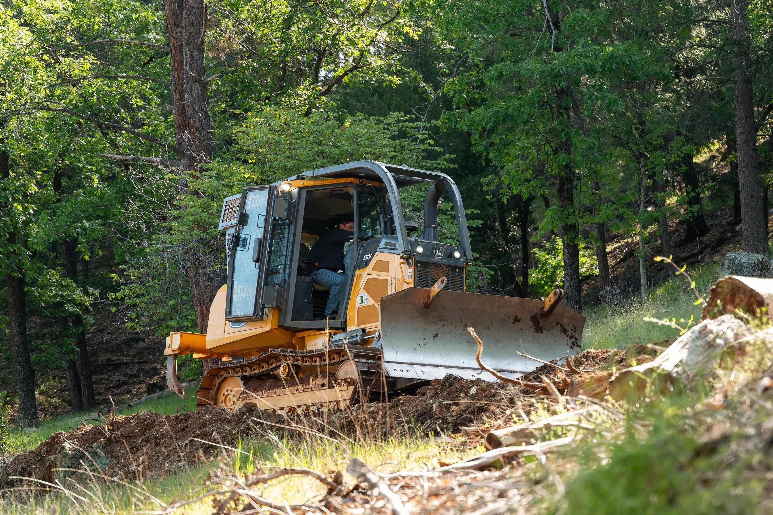 A person operating a yellow bulldozer in a forested area with green trees and logs on the ground.