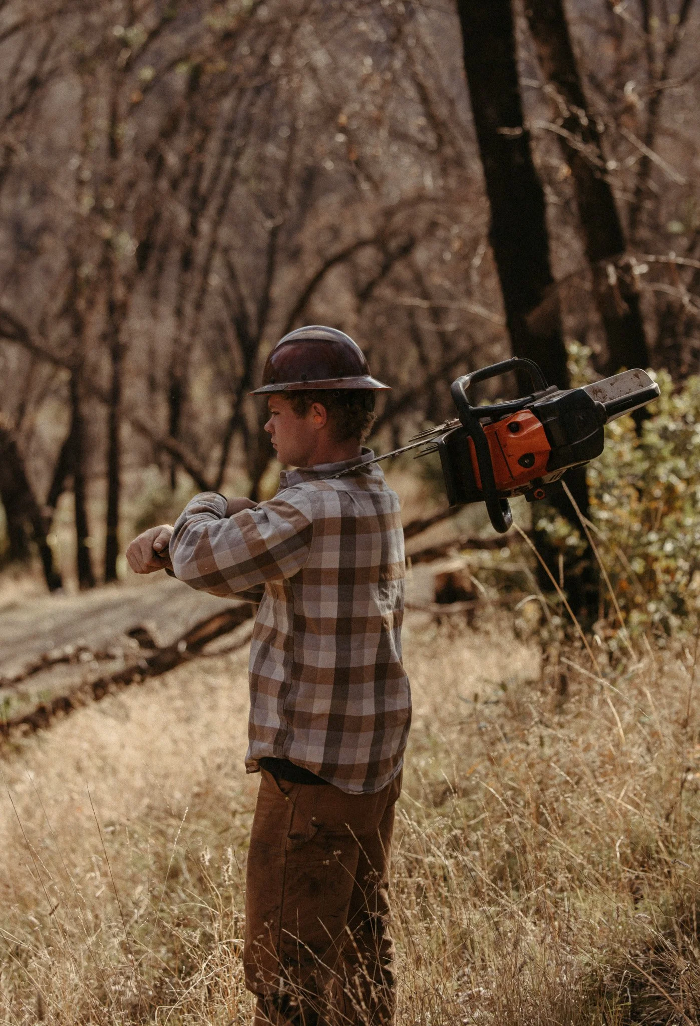 A young boy wearing a hard hat and plaid shirt standing outdoors in a wooded area, preparing to use a chainsaw.