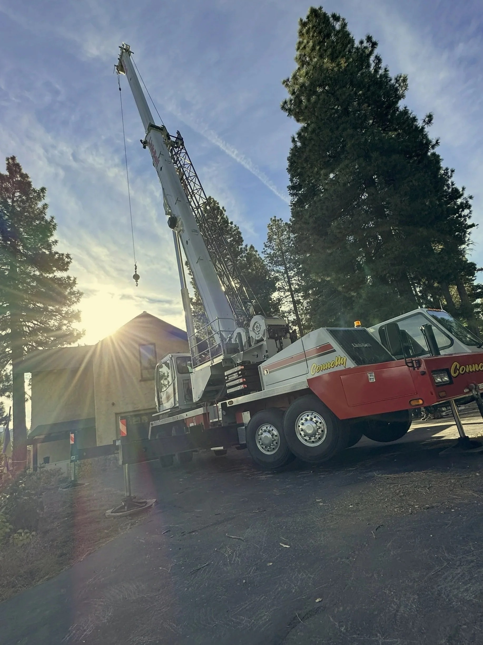 A large white and red crane truck parked on a paved surface near a beige building surrounded by tall trees, with the sun setting behind the building and trees.